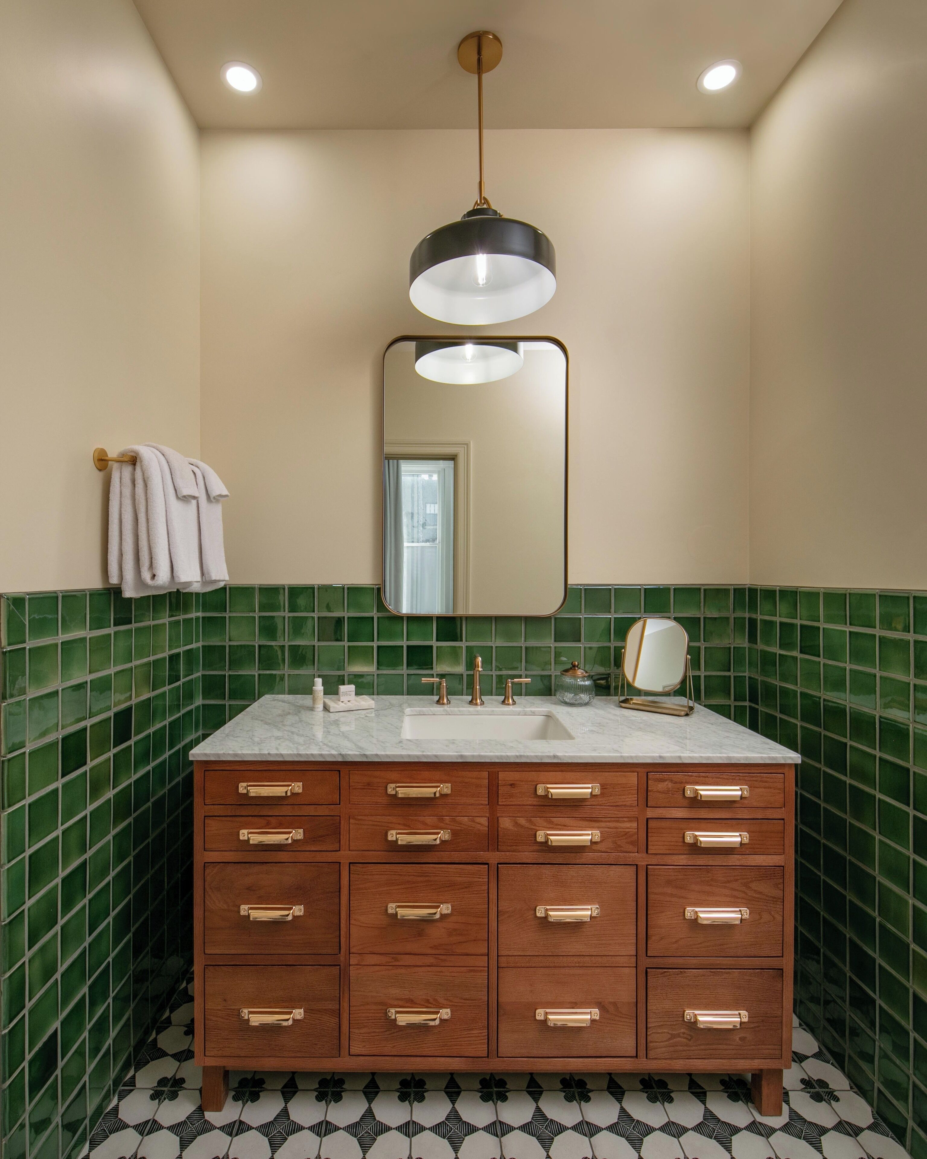 A bathroom with a wooden vanity, green tiled walls, a mirror, and a hanging light fixture.