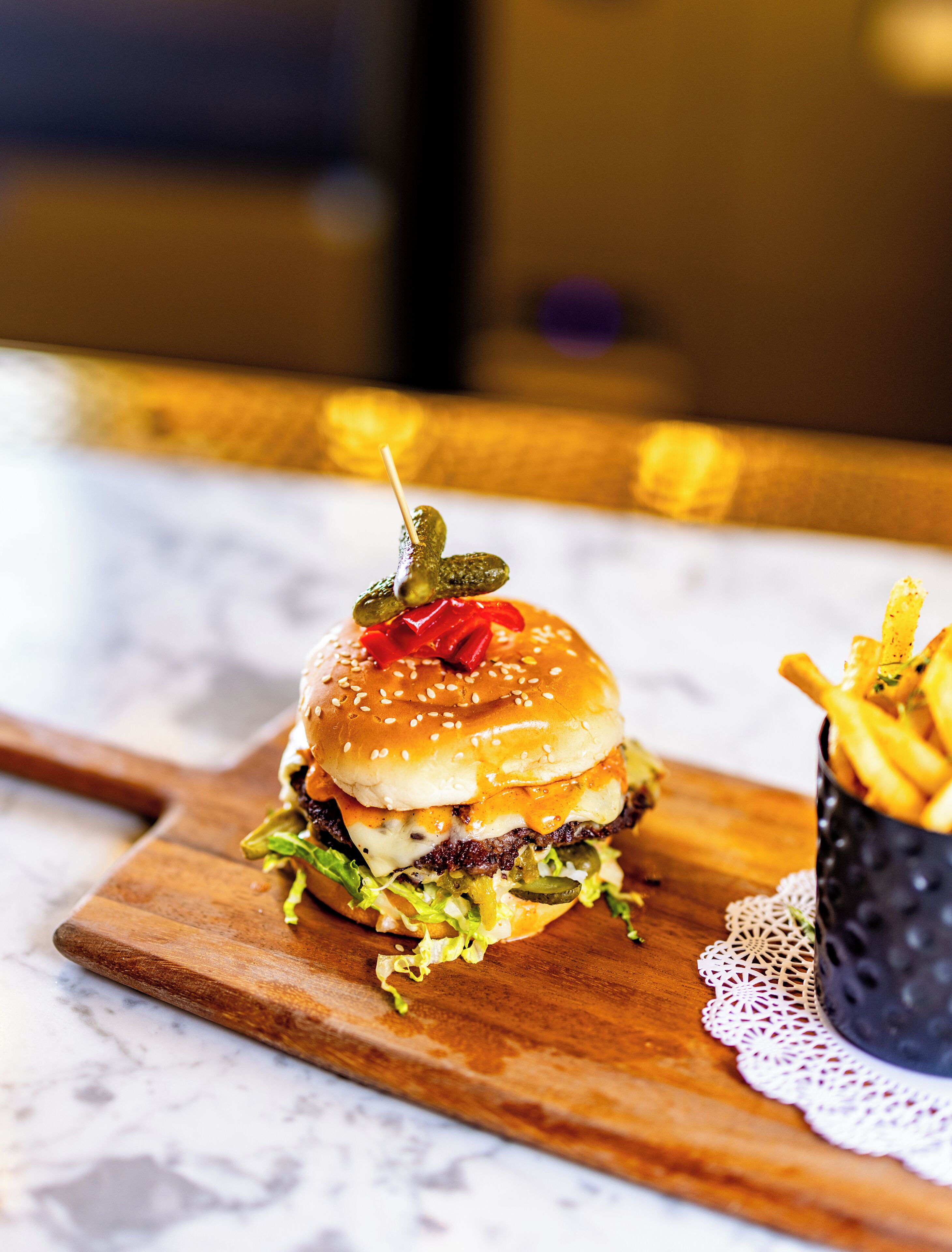 A burger with pickles and a red pepper on a sesame seed bun, served with fries on a wooden board.