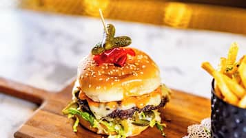 En burger med syltede agurker og en rød peber på en sesambrød, serveret med pommes frites på en træplanke.