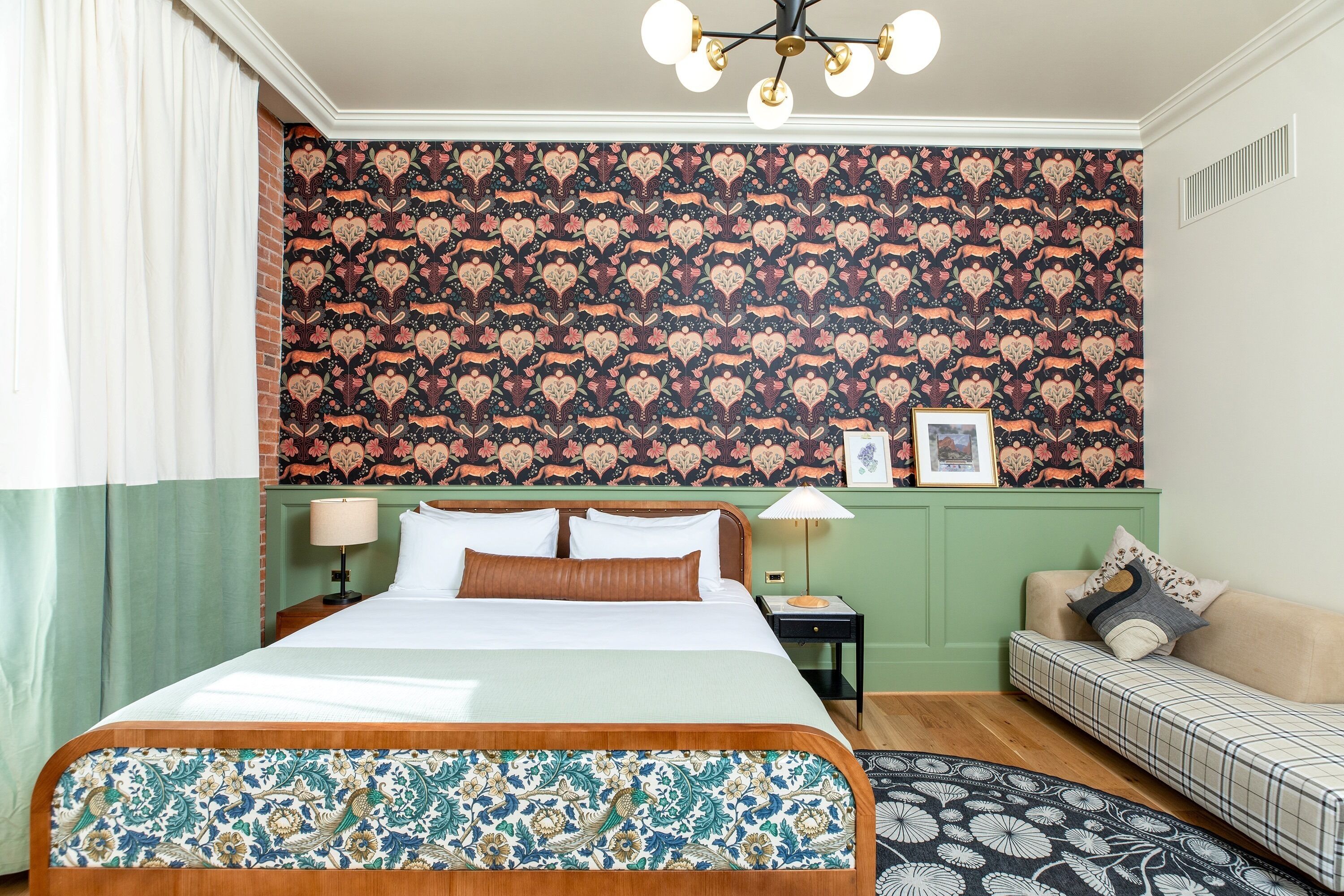 A hotel room with a large bed, a green accent wall, a patterned sofa, and a chandelier.