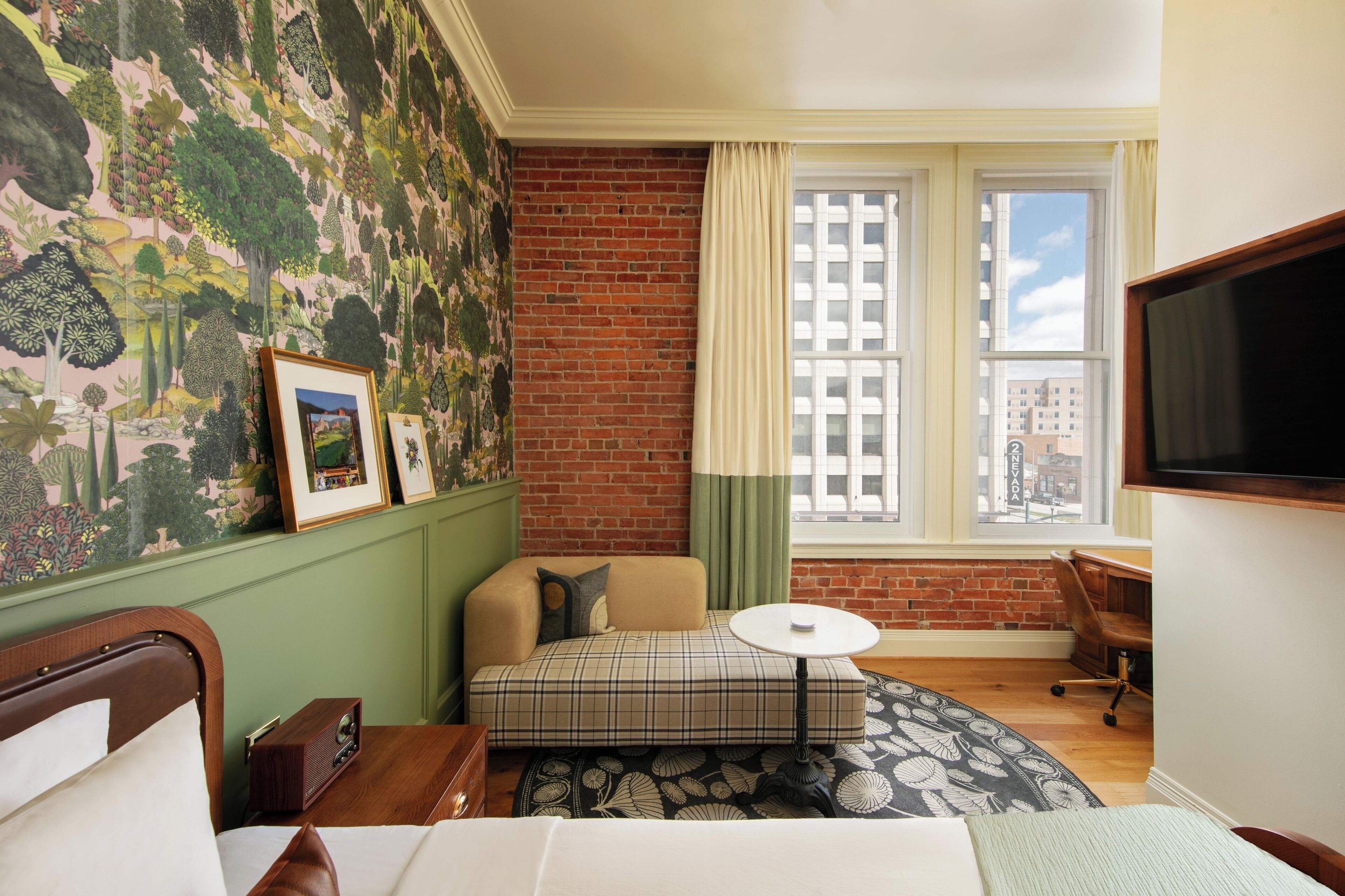 Hotelkamer met een groene wand, een accentwand van baksteen, een groot raam met uitzicht over de stad, een bed met houten hoofdeinde, een kleine bank, een ronde tafel en een televisie aan de wand.
