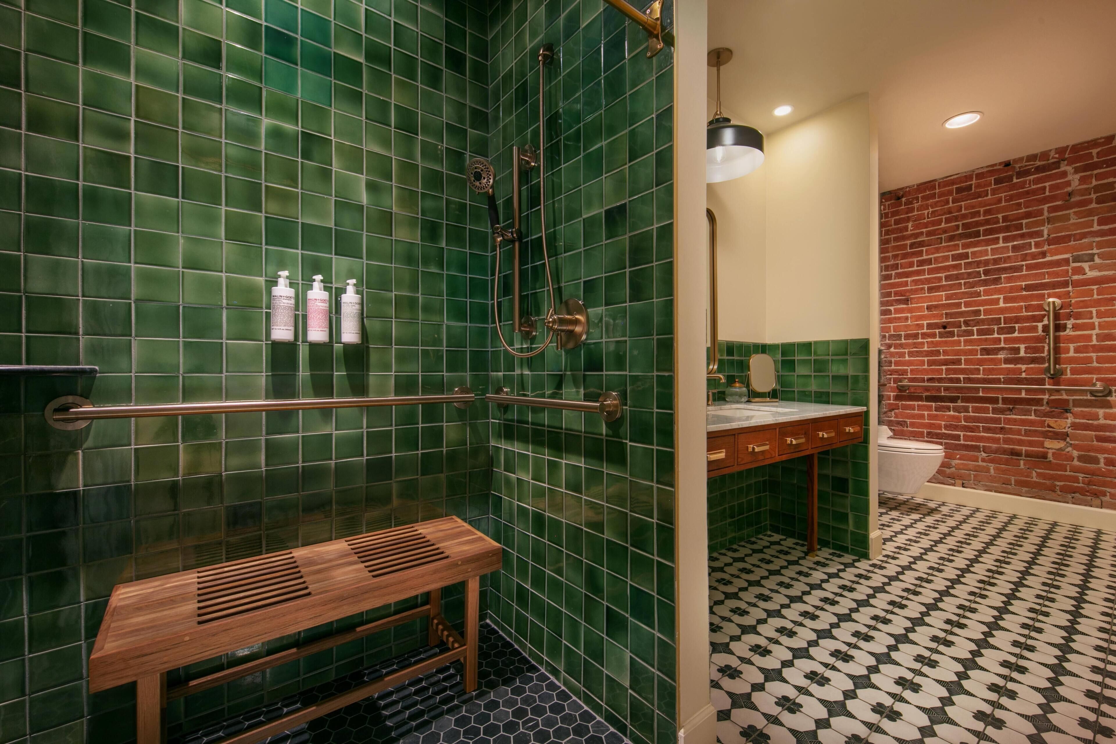 A bathroom with green tiled walls, a wooden bench, a shower with a handheld showerhead, and a toilet.
