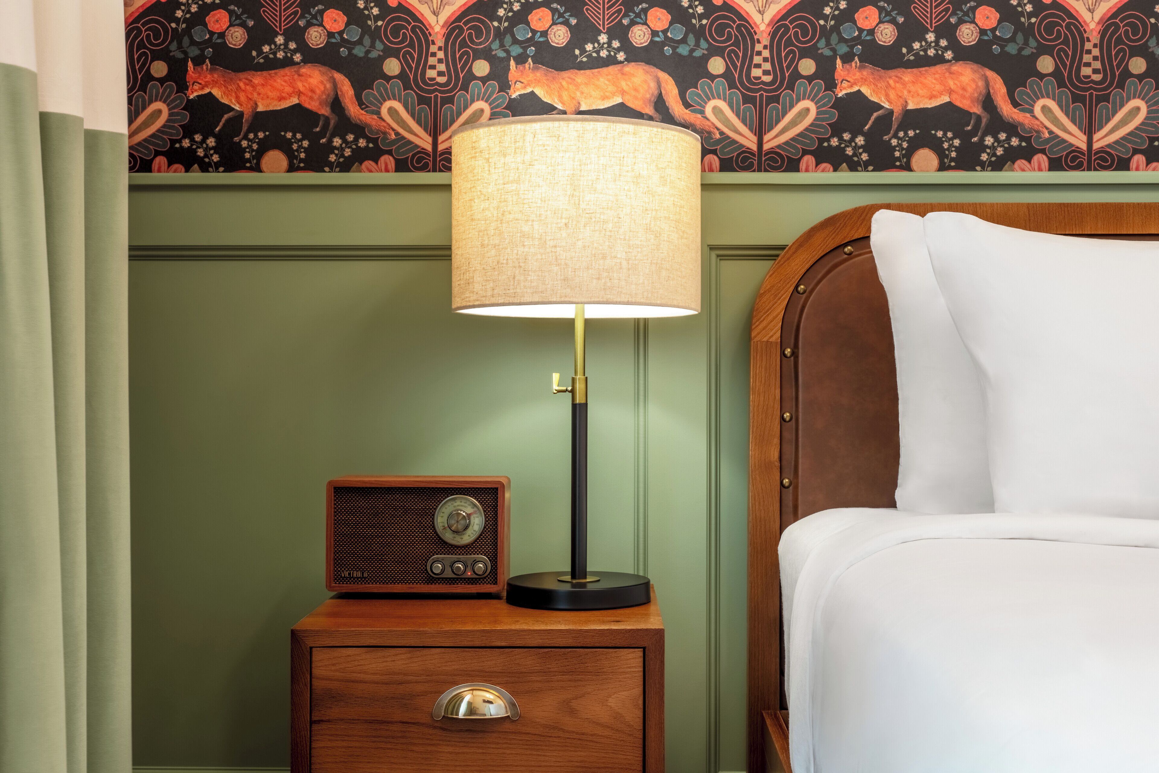Una mesita de noche de madera con una radio y una lámpara, una cama con sábanas blancas y una pared con papel tapiz estampado con zorros.