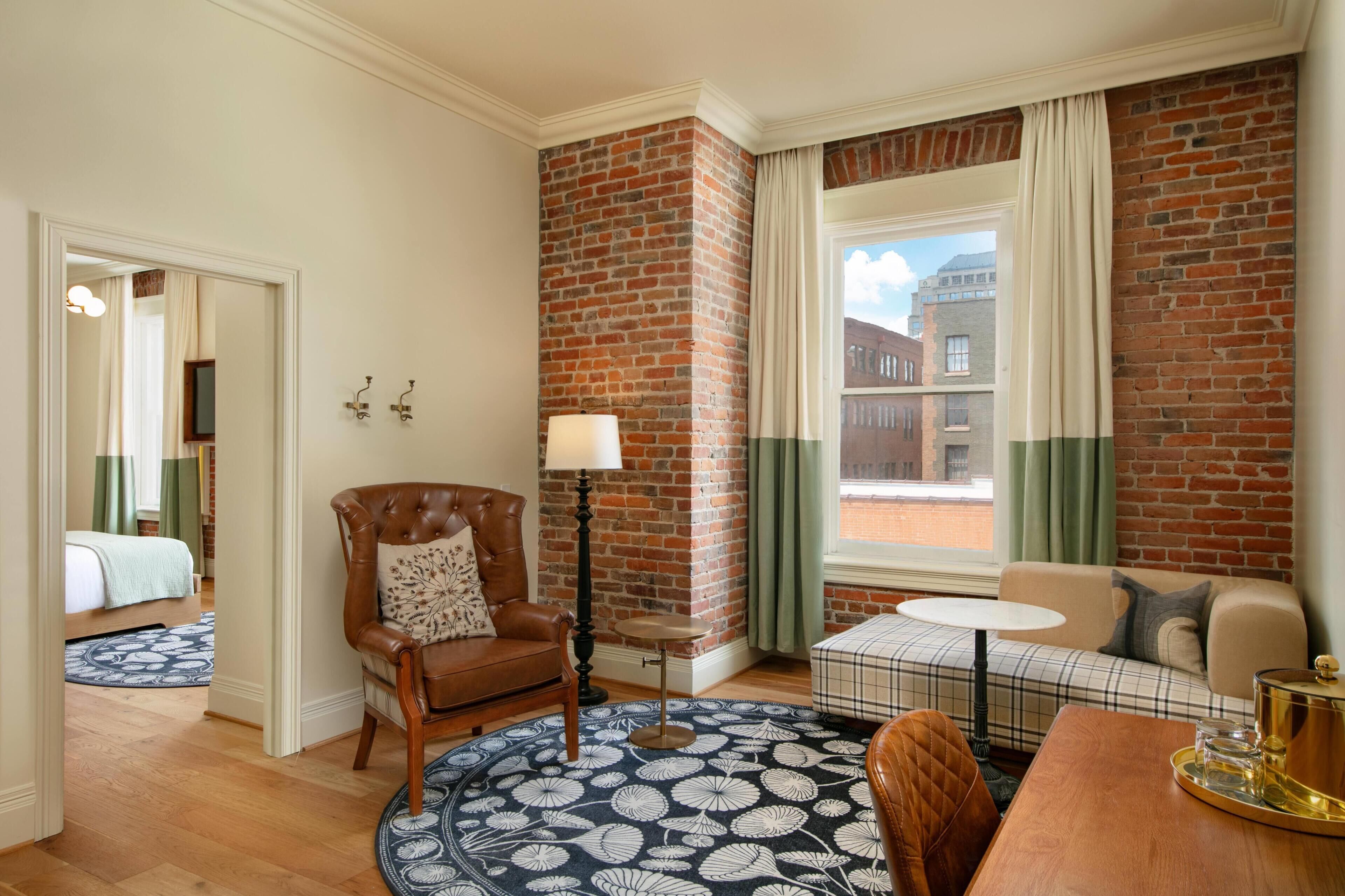 A hotel room with a brick accent wall, a round rug, a sofa, a chair, a small table, and a window with curtains.
