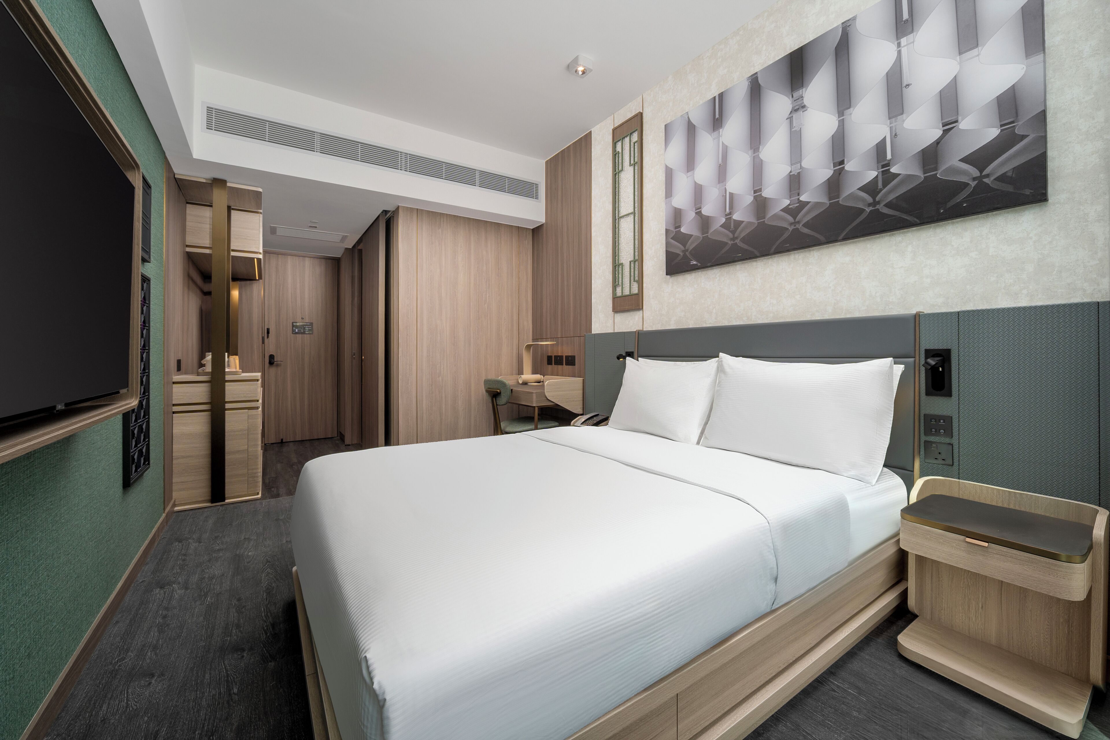 A modern hotel room with a large bed, a desk, a chair, a TV, and a wall-mounted artwork.