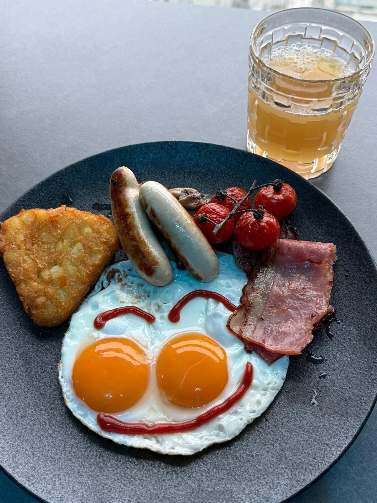A plate of breakfast food including eggs, sausages, bacon, and tomatoes, with a glass of beer in the background.