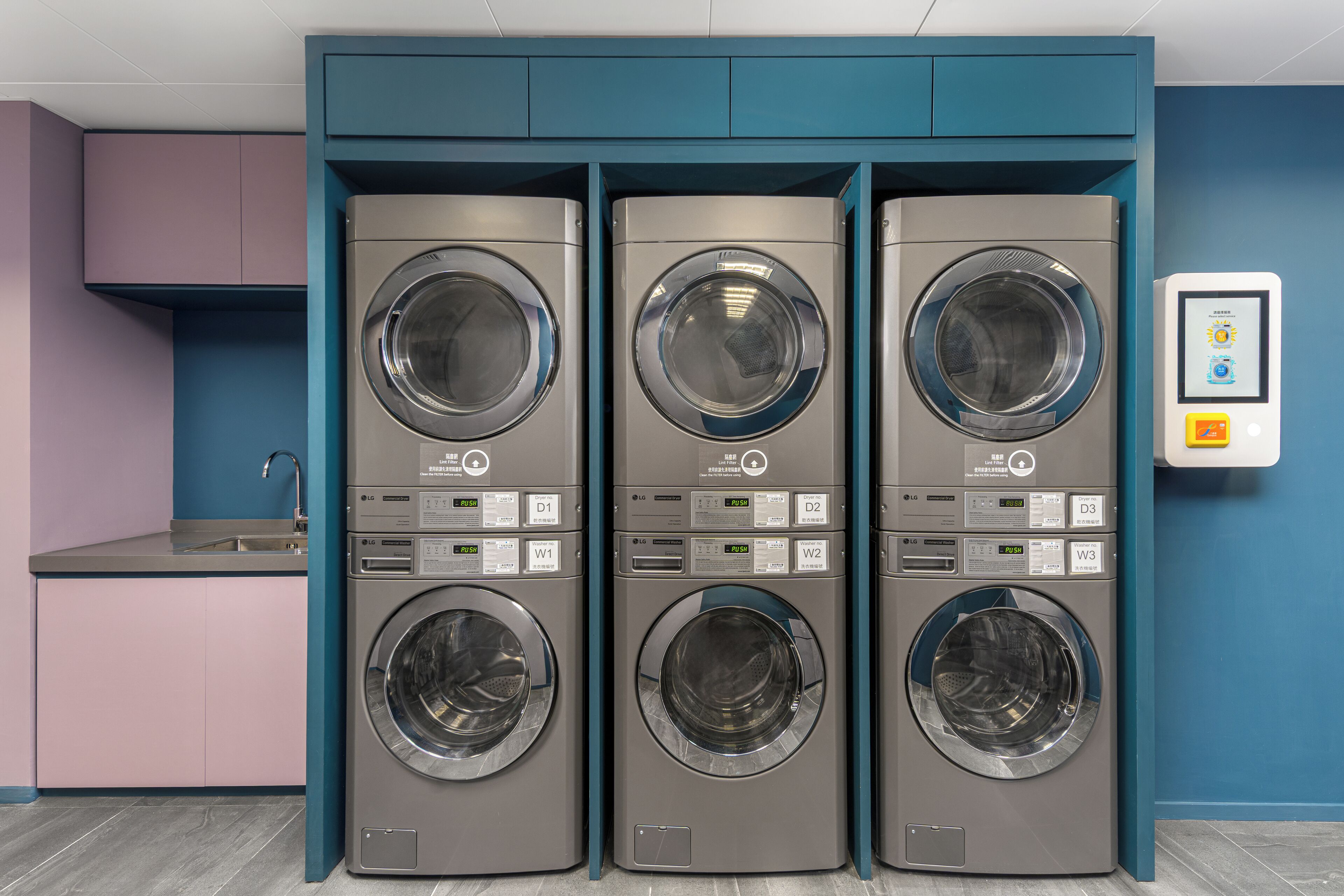 Three stacked front-loading washing machines in a modern laundry room with teal cabinetry and a pink countertop.