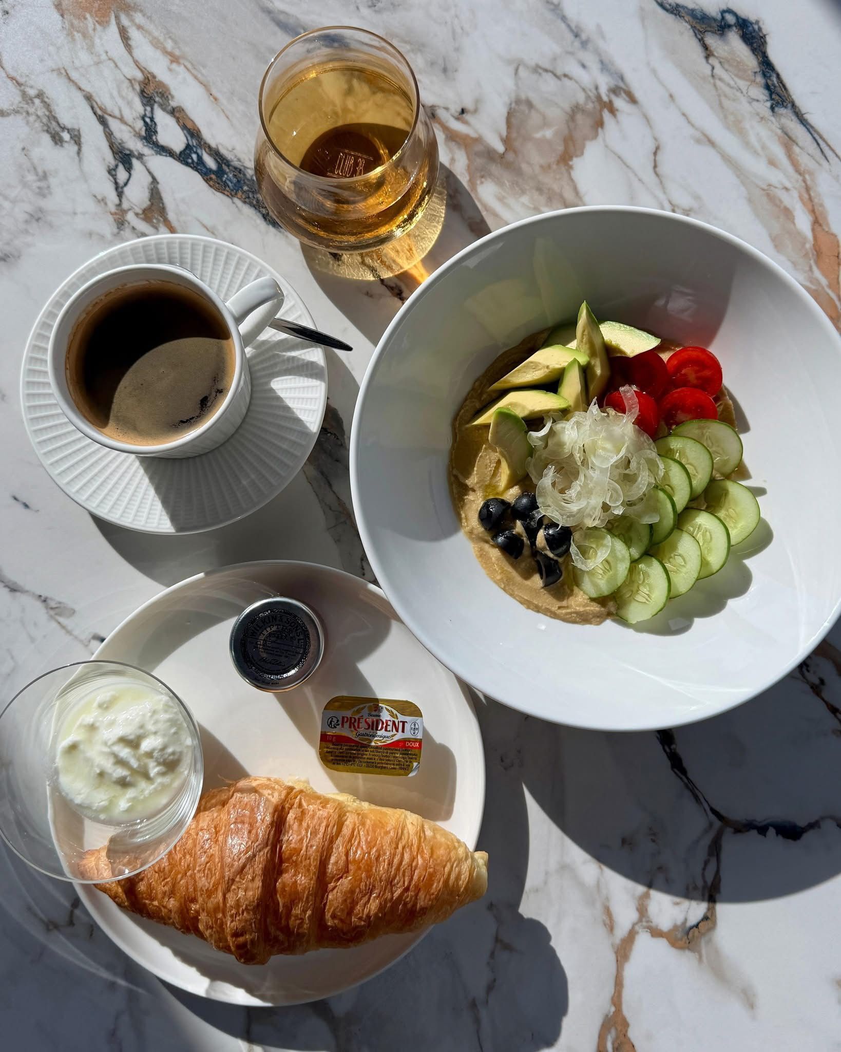 A breakfast setting with a croissant, a bowl of fruit salad, a cup of coffee, and a glass of juice on a marble table.