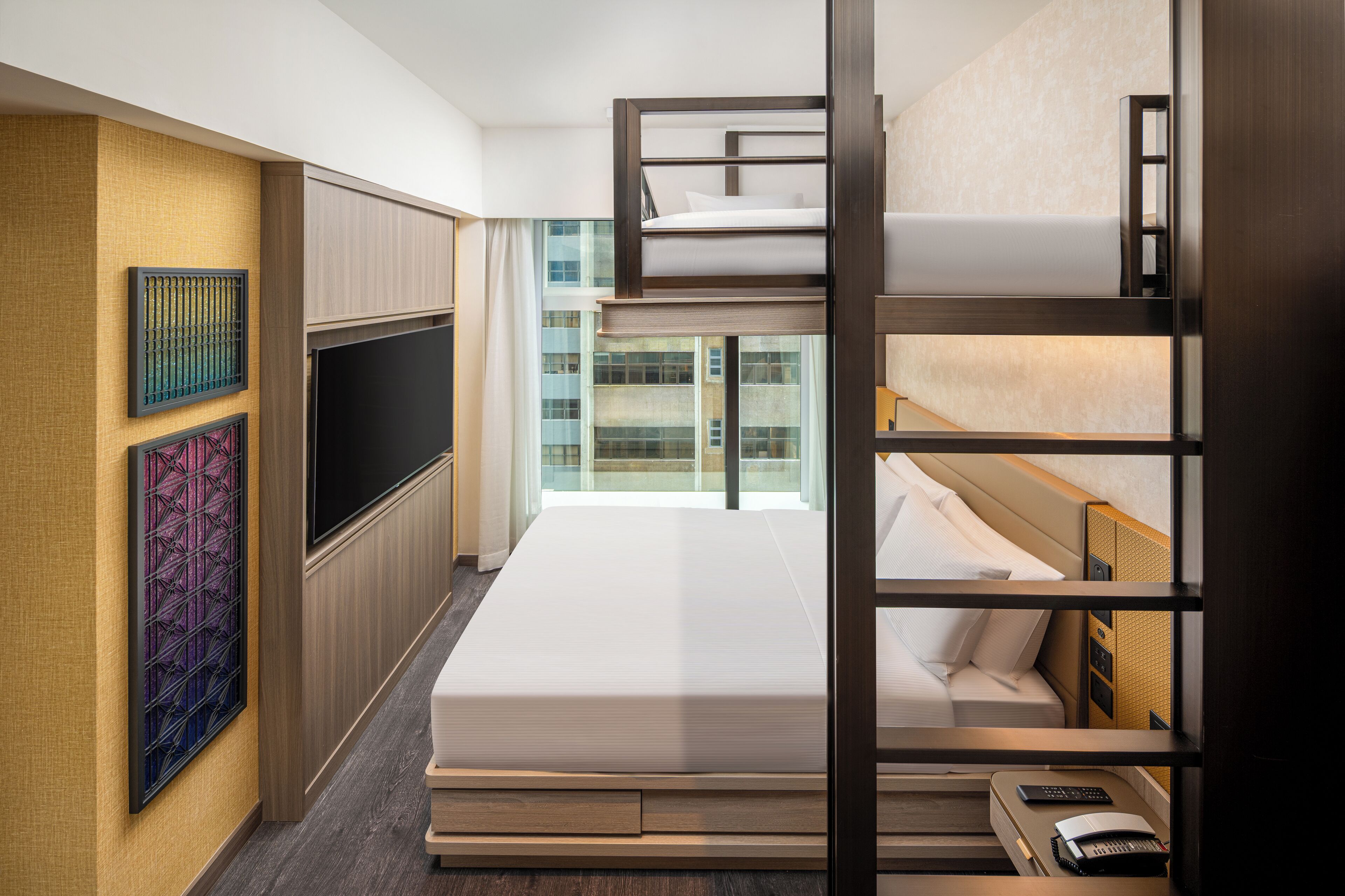 A hotel room with a bunk bed, a flat-screen TV, a desk with a telephone, and a window with a city view.