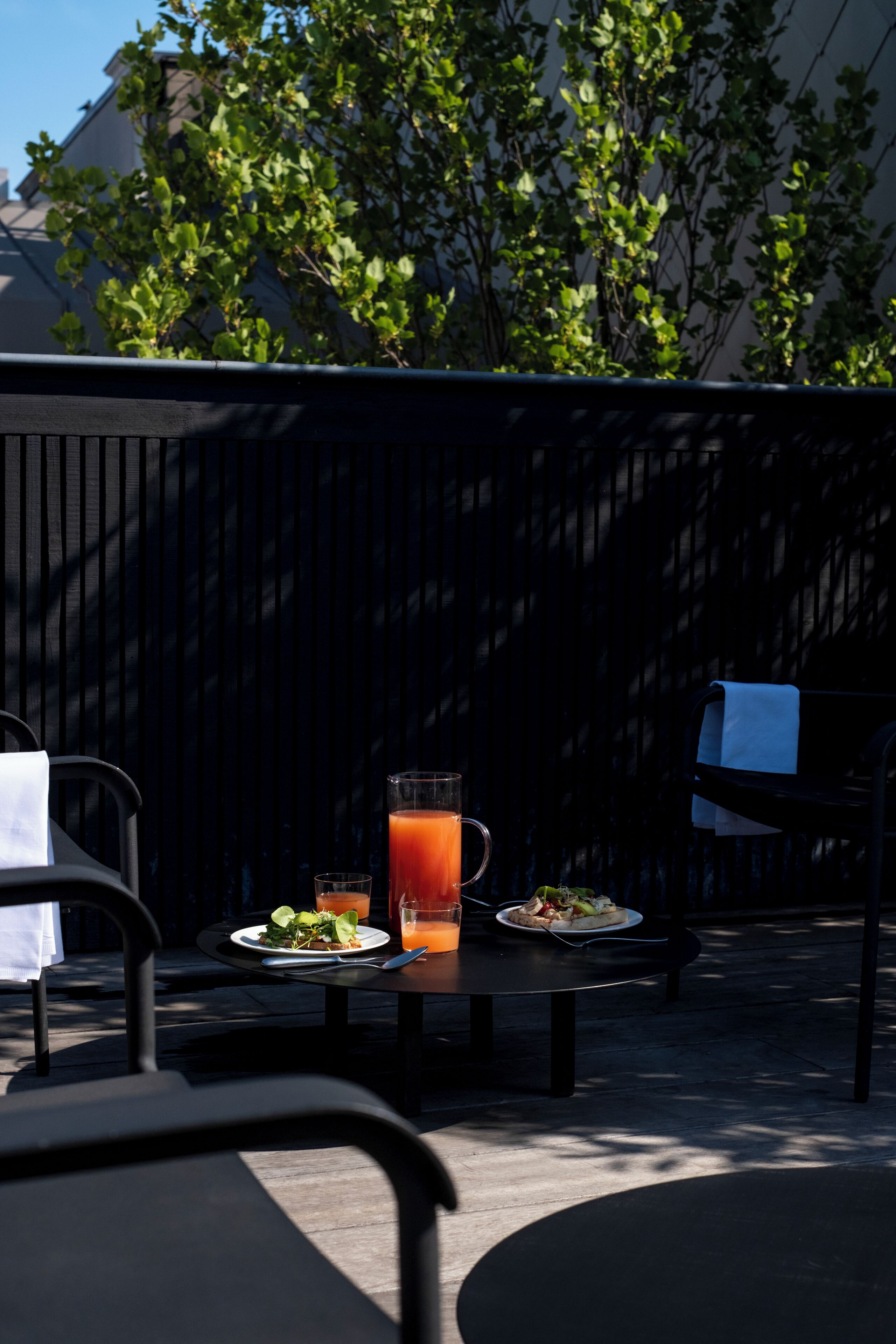 Un espace de restauration en plein air avec une table dressée pour deux personnes, une carafe de thé glacé et une assiette de nourriture.