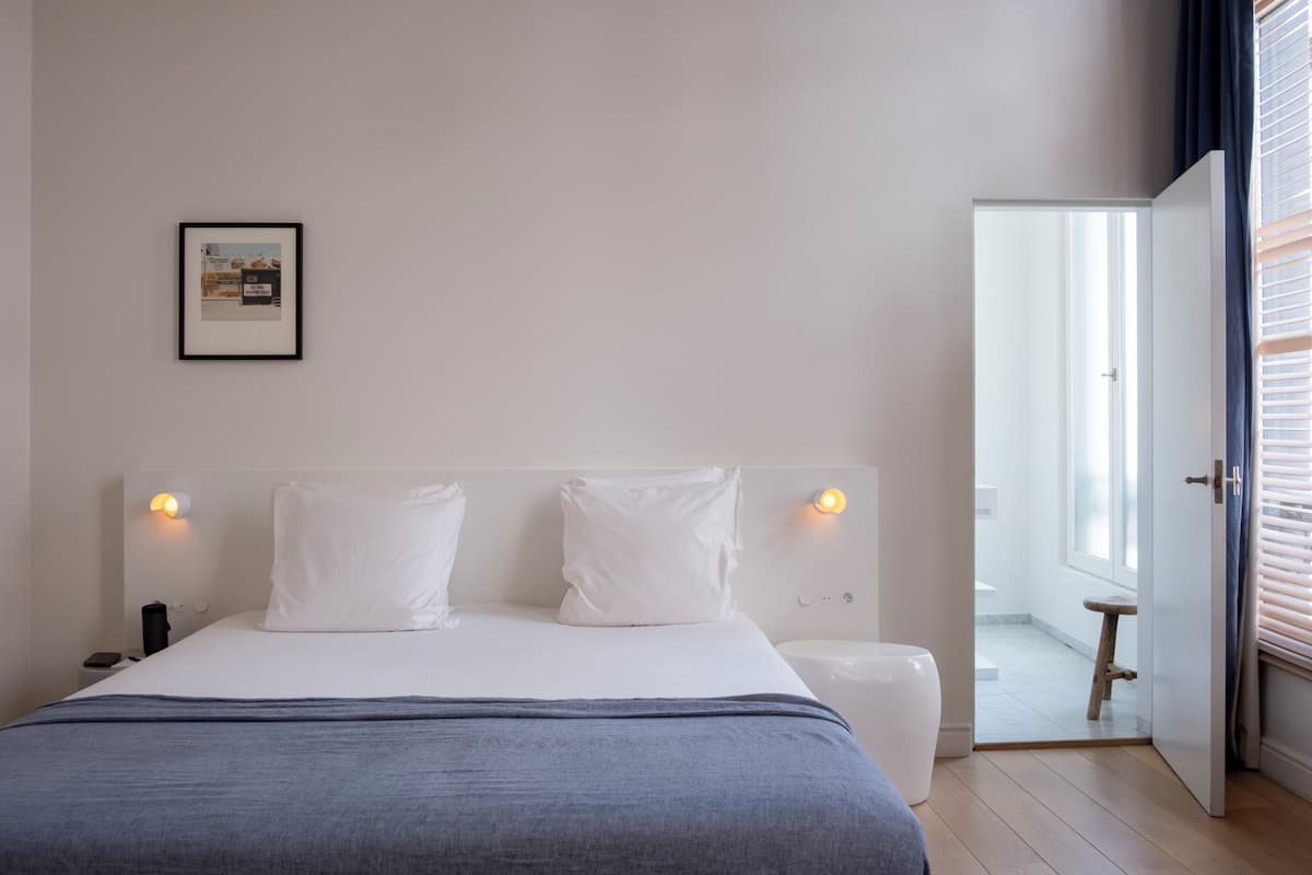 Une chambre moderne avec un grand lit, des lampes de chevet, un tableau encadré au mur et une vue sur la salle de bain par une porte ouverte.