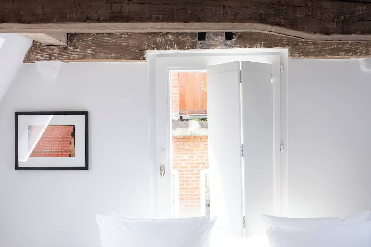 Une chambre à coucher avec un lit, des oreillers, une table de chevet, un tableau encadré et une fenêtre donnant sur un immeuble en briques.