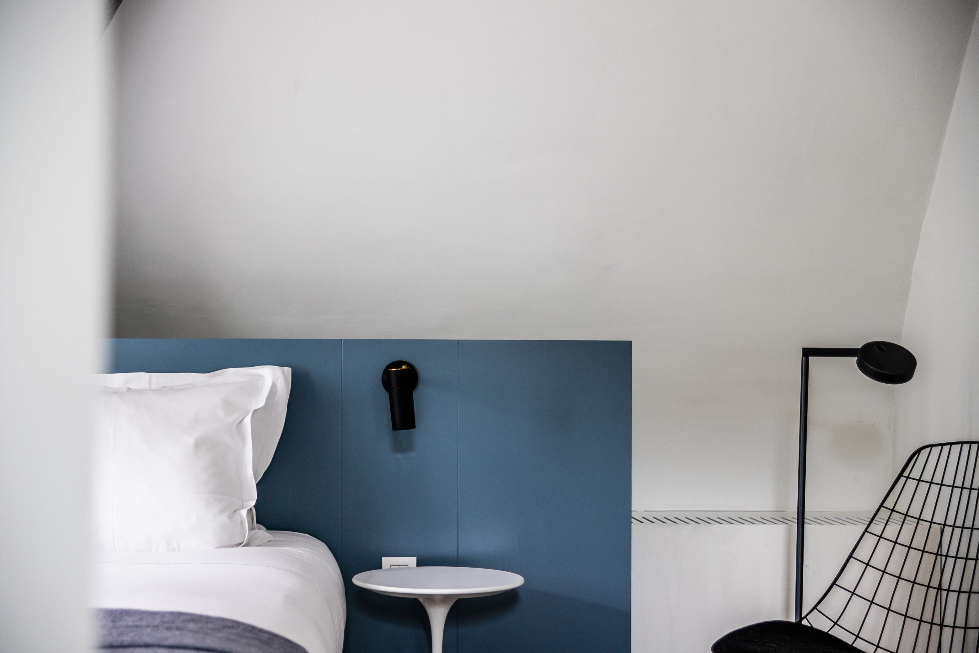 A modern hotel room with a bed, a small round table, a chair, a wall-mounted lamp, and a radiator.