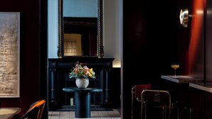 A dimly lit hallway with a wooden floor, a black table with a vase of flowers, and a bar area with stools and a bottle of wine. - Hotel Julien (Antwerp)