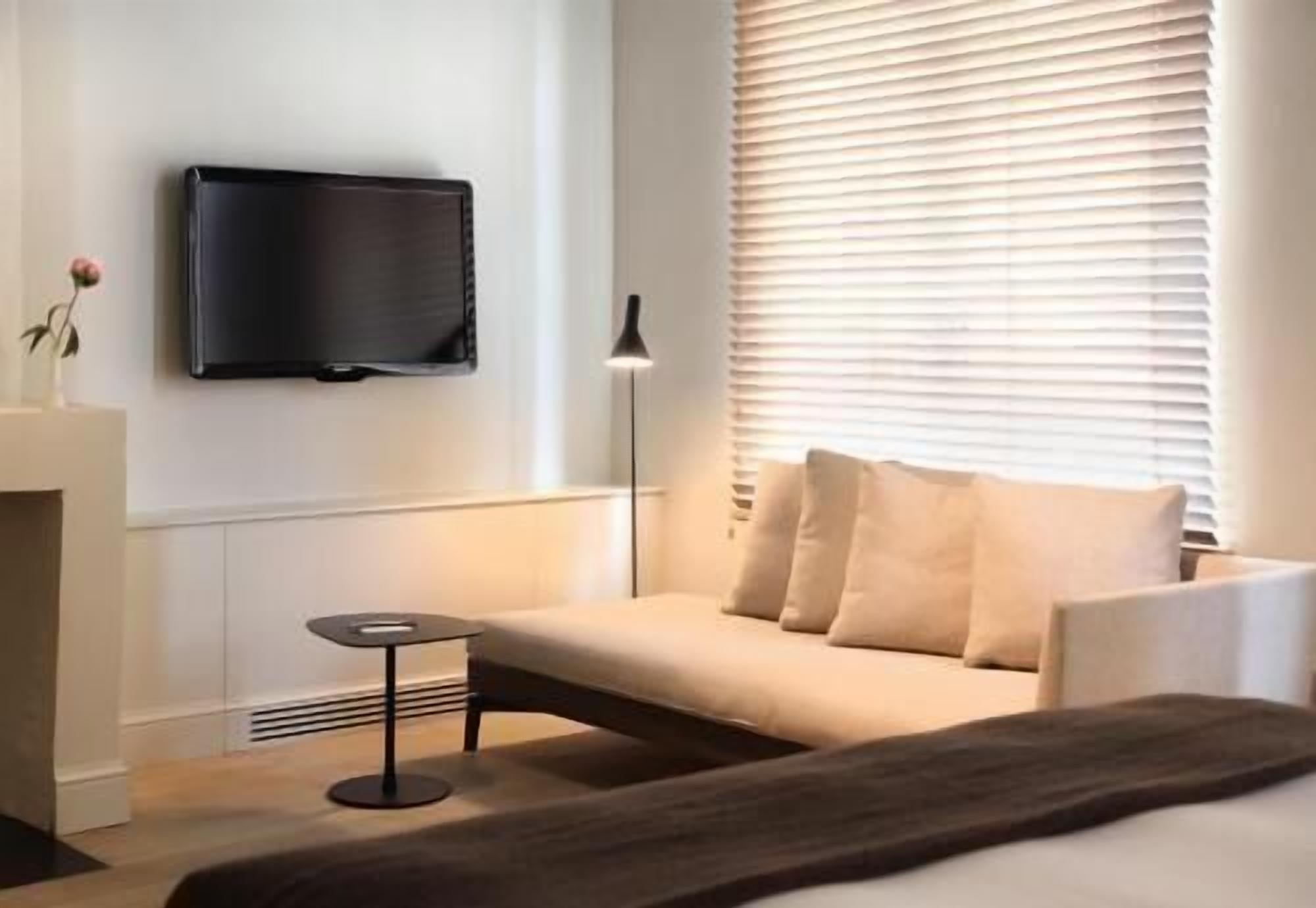 A modern hotel room with a flat-screen TV mounted on the wall, a sofa with cushions, a small round table, and a floor lamp.