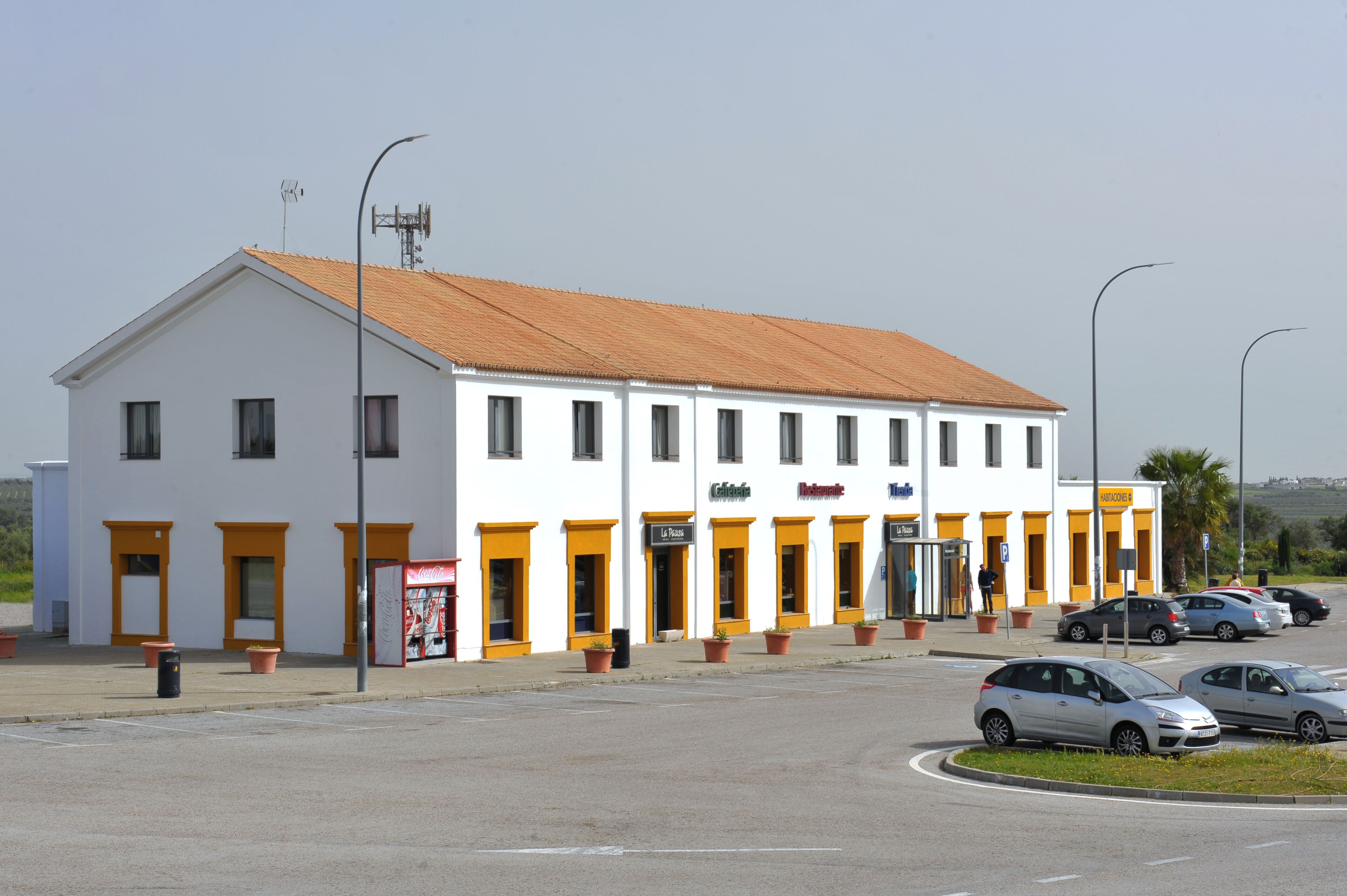 Un edificio blanco con detalles naranja, que cuenta con varias puertas y ventanas, y un letrero con distintos nombres de negocios.