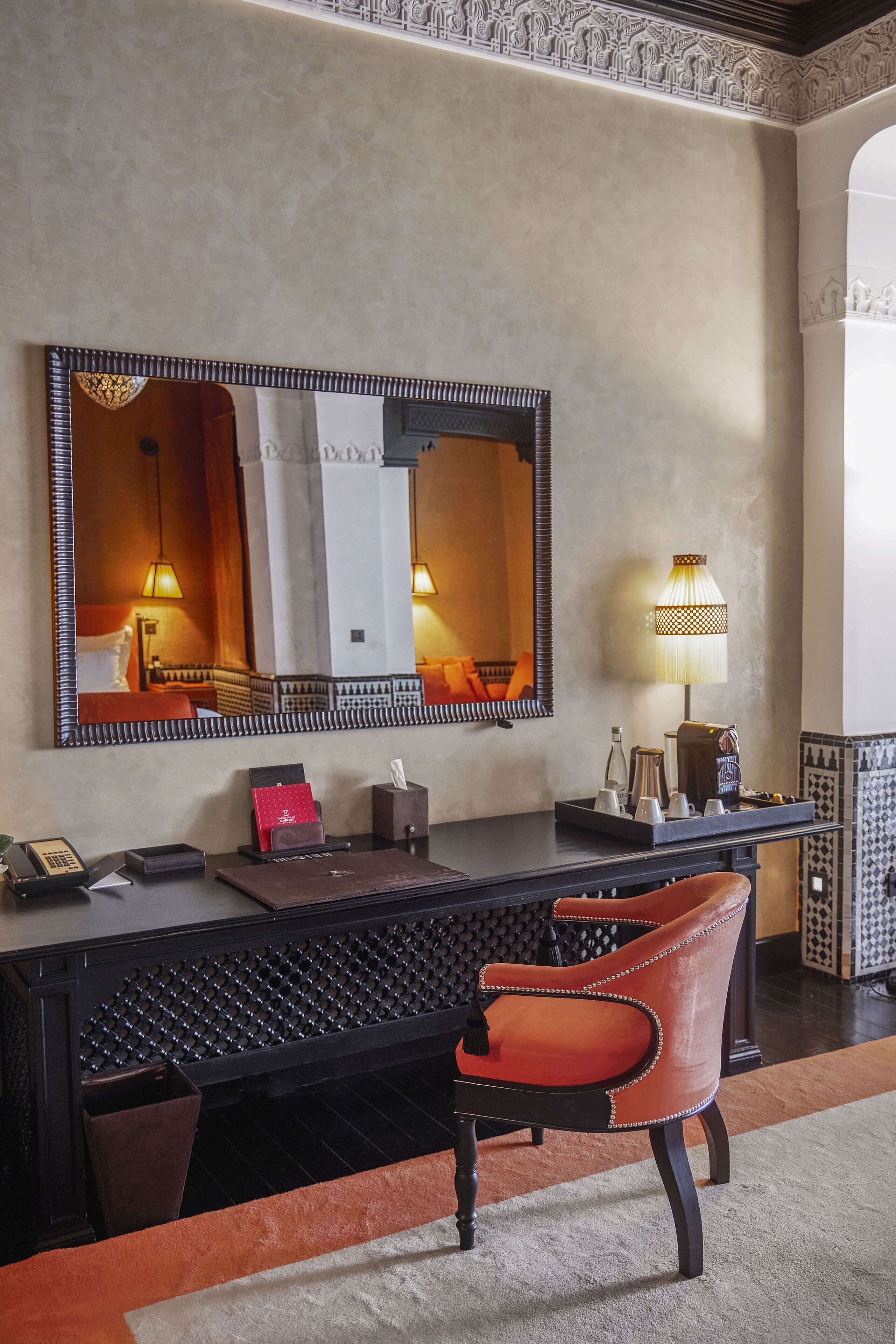 Una habitación de hotel con un espejo grande, un escritorio con teléfono, una lámpara y una silla.