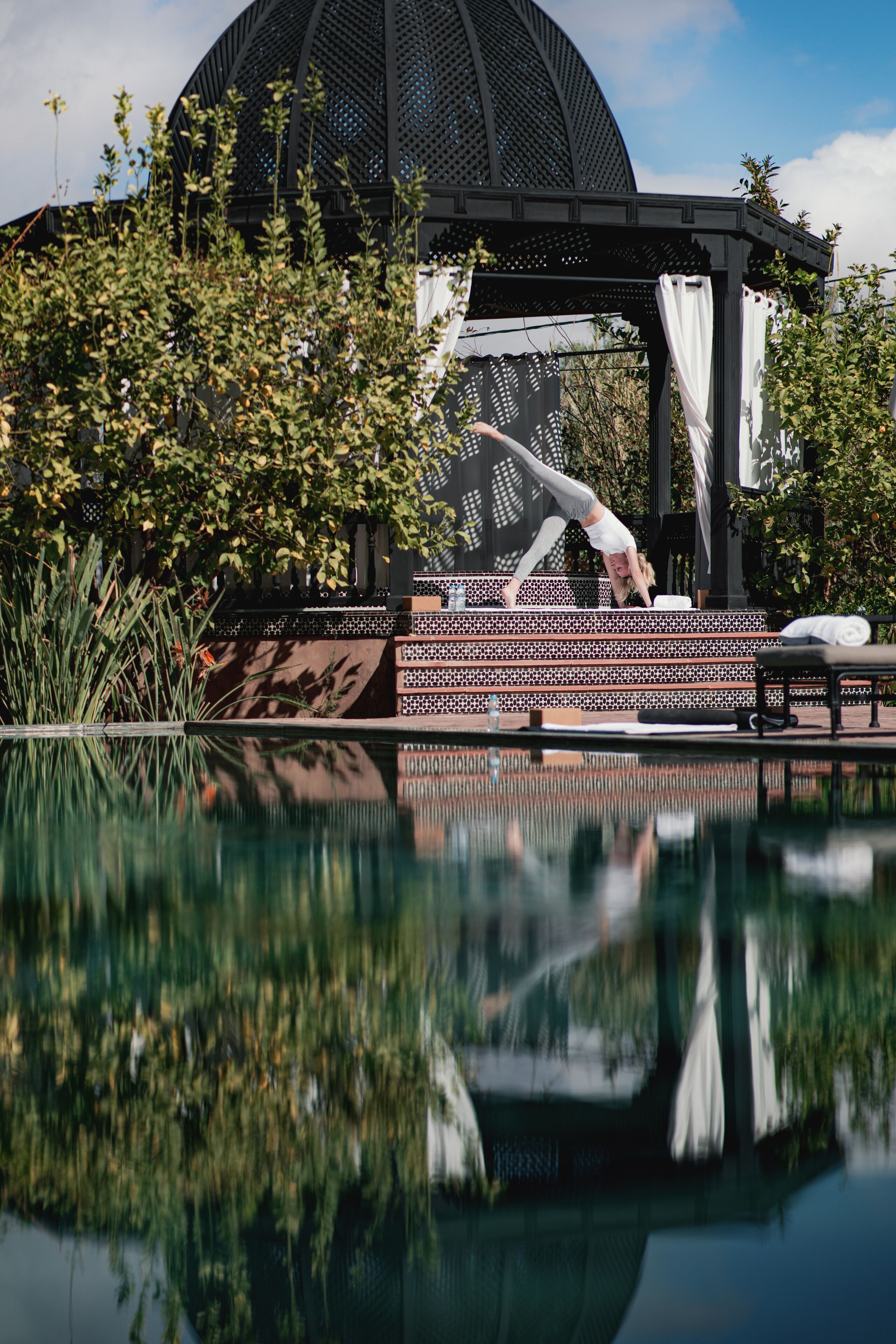 A person performing a yoga pose on a platform by a reflective pool with a gazebo and greenery in the background.