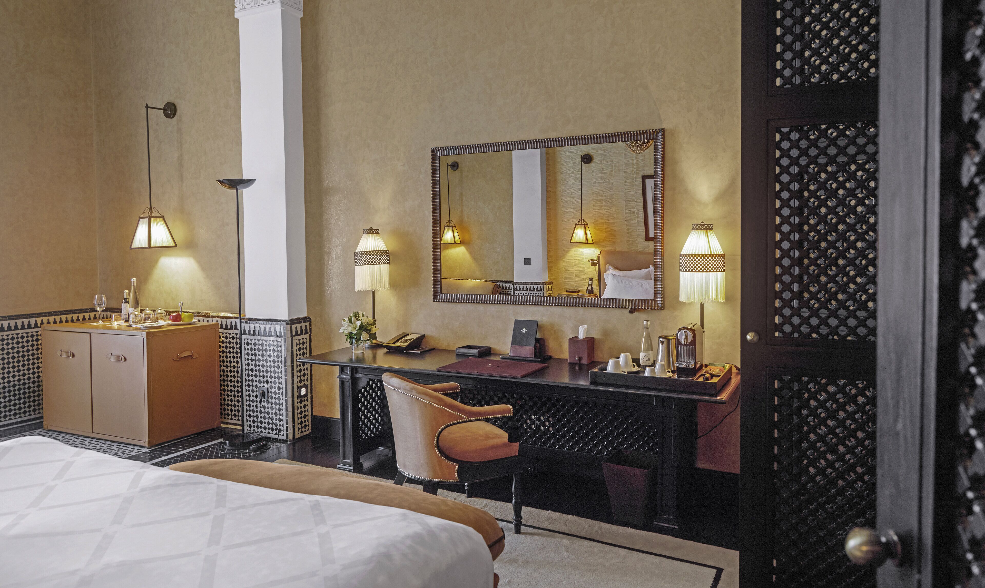 A hotel room with a bed, a desk with a telephone, a chair, a minibar, and a large mirror.