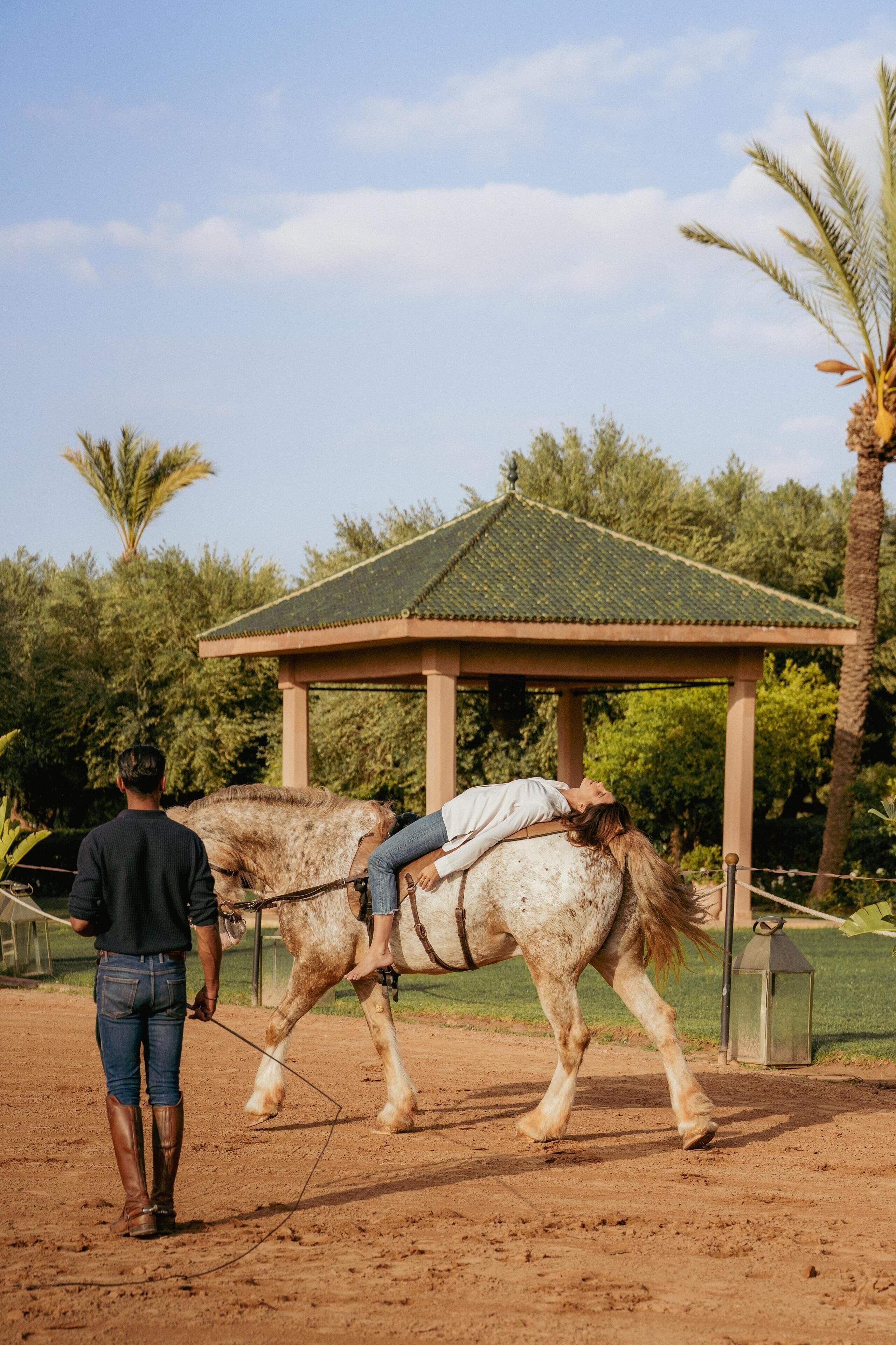 A person leading a horse with a saddle, walking on a dirt path with a gazebo in the background.