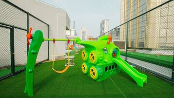 A playground with a slide and climbing structures, surrounded by a chain-link fence and artificial grass.