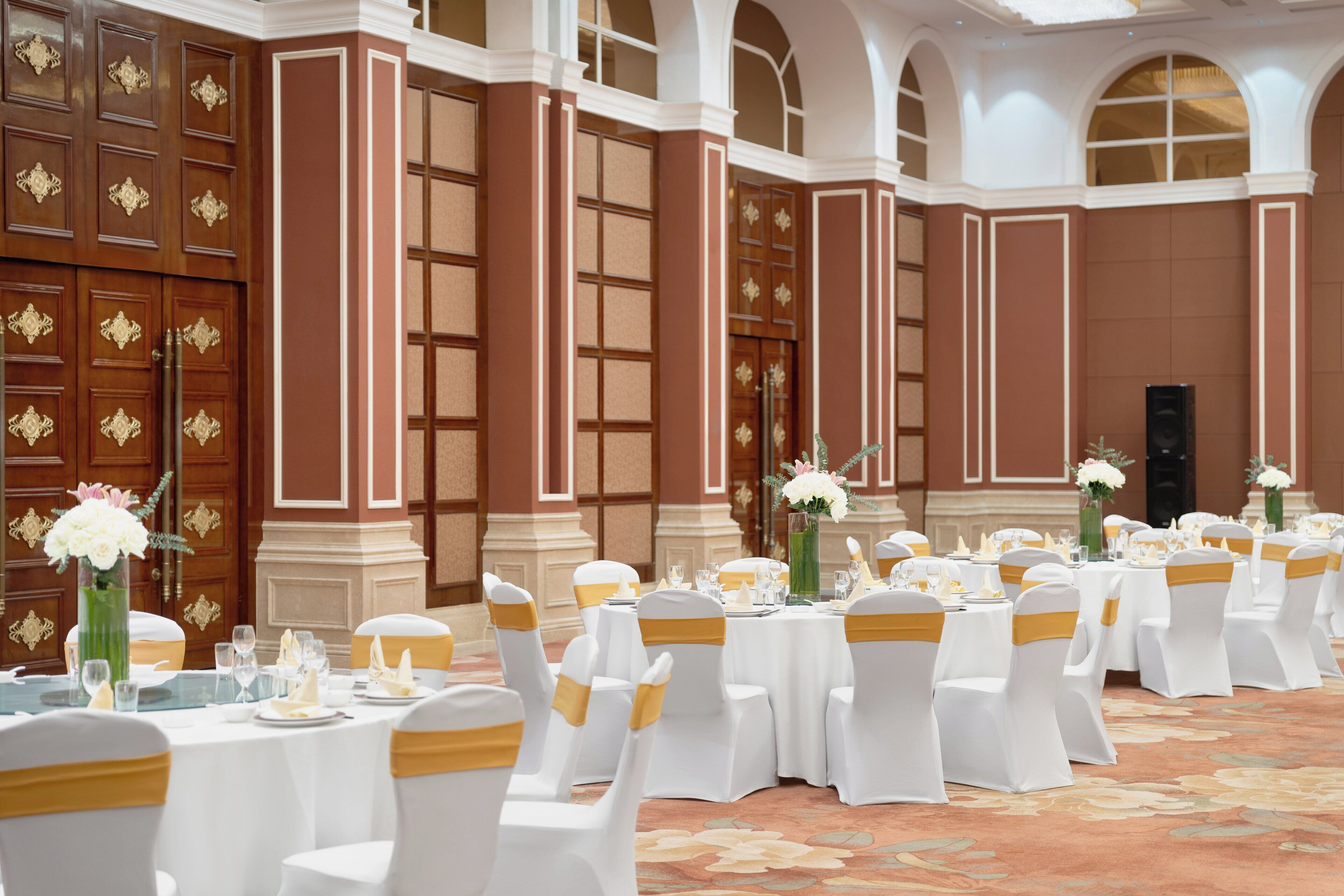 A banquet hall with round tables set for a formal event, featuring white chairs with gold sashes, floral centerpieces, and a classic architectural design with arched windows and detailed wood paneling.