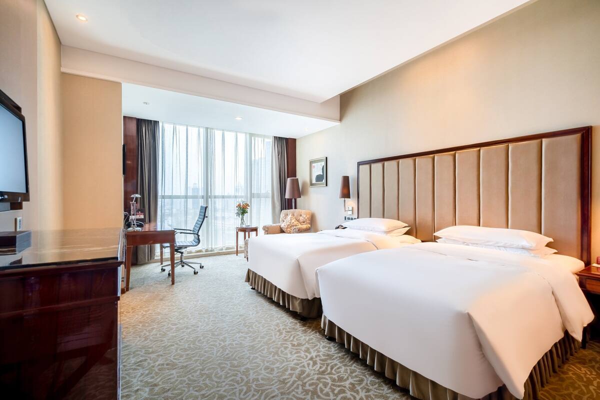 Habitación de hotel con dos camas, un escritorio, una silla, un televisor y vistas de la ciudad a través de amplias ventanas.