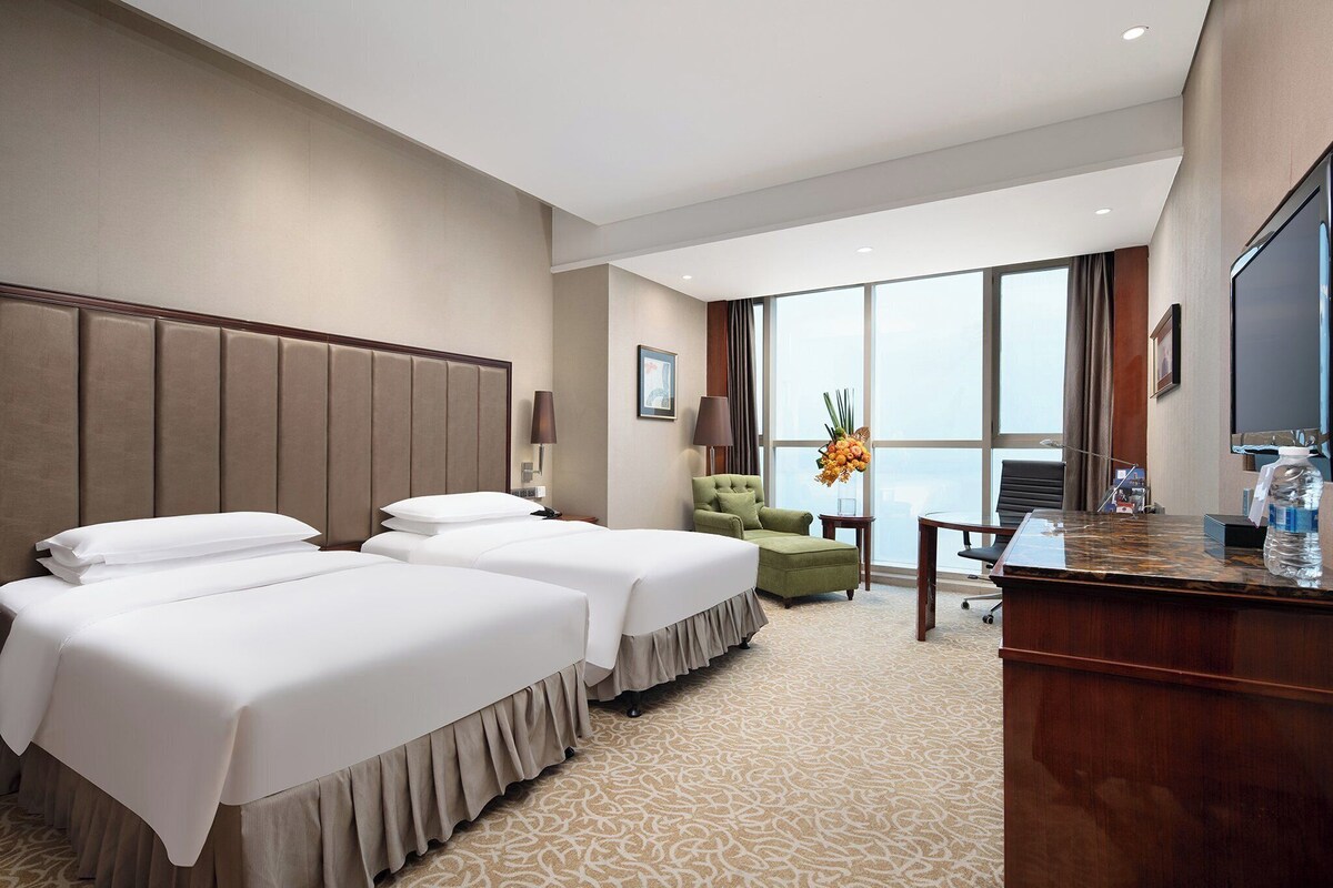 A hotel room with two beds, a desk, a chair, a TV, and a large window.