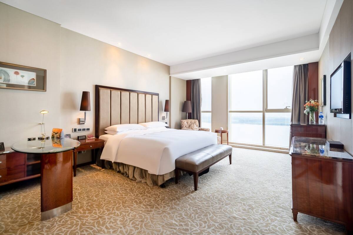 Habitación de hotel con una cama grande, un escritorio con lámpara, una silla, un televisor y un ventanal con vistas.