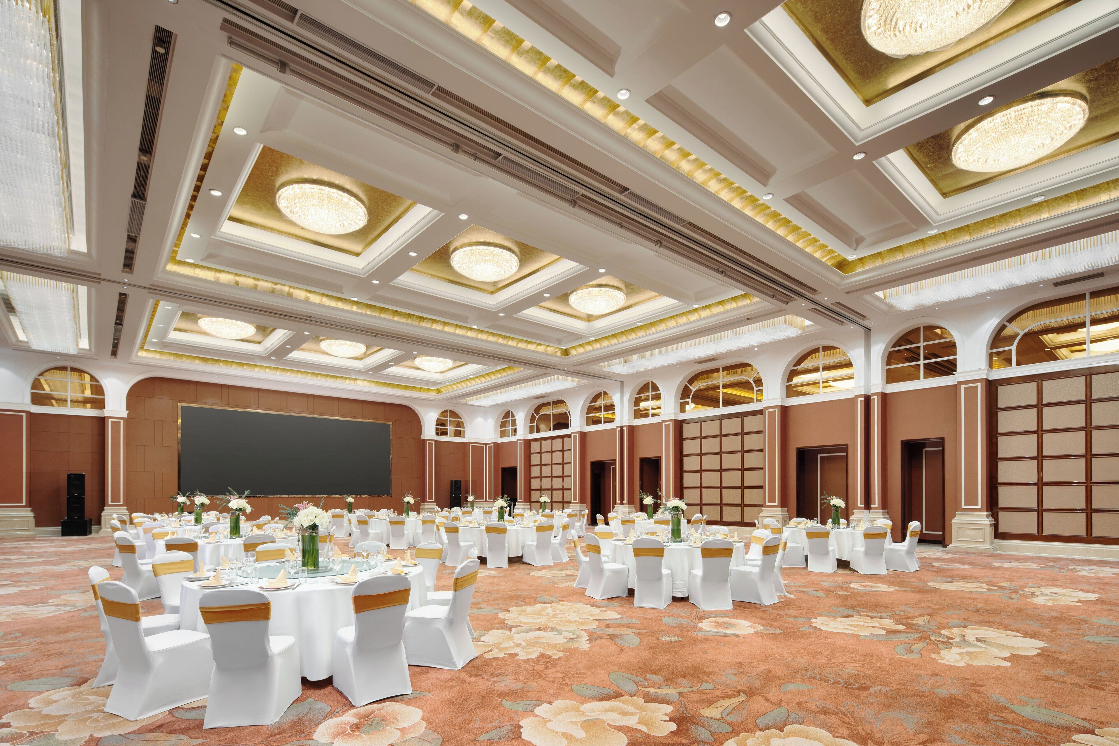 A large conference room with multiple round tables, white chairs, and floral carpet.