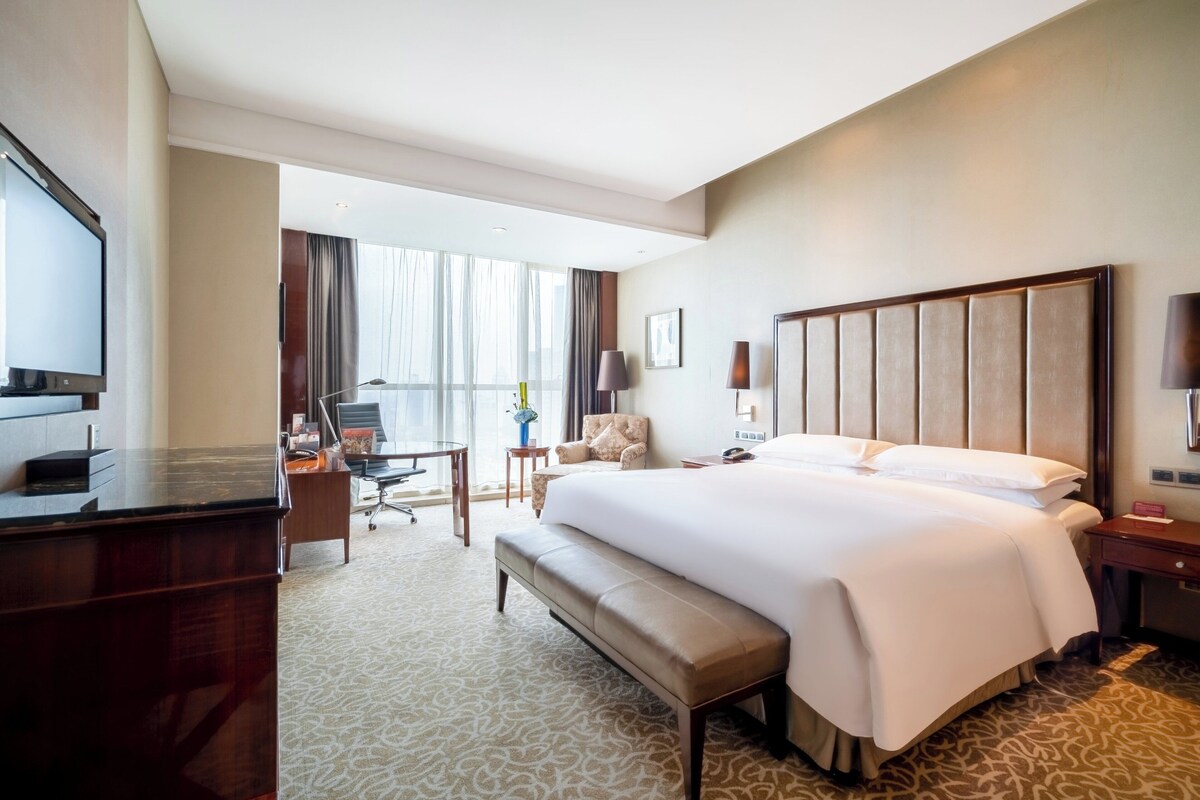 A hotel room with a large bed, a desk, a chair, a television, and a window with curtains.