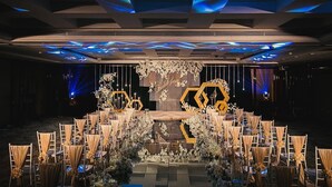 Un escenario con un arreglo floral geométrico y una estructura hexagonal, rodeado de filas de sillas con manteles.