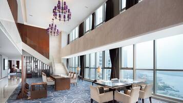 A spacious hotel lobby with a high ceiling, a grand staircase, a dining area with a glass table, and large windows offering a city view.