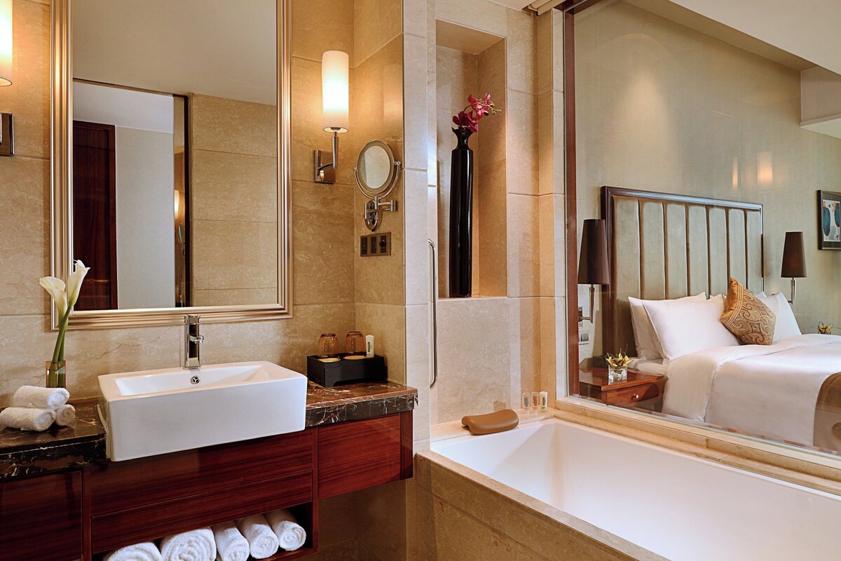 A hotel bathroom with a large mirror, a sink, a bathtub, and a bed visible in the reflection.