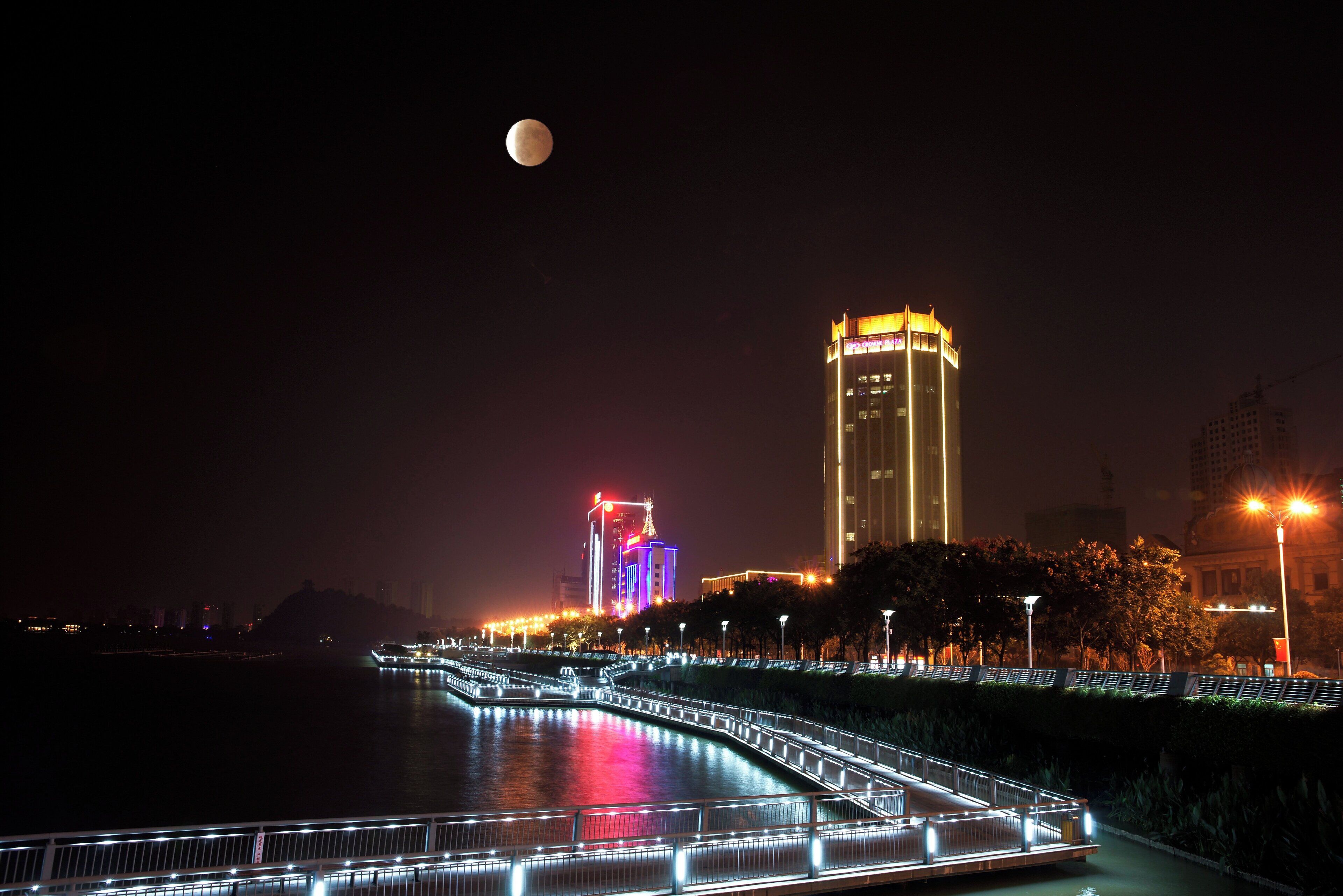 Une vue nocturne d’une ville, avec une rivière, un pont illuminé et un bâtiment imposant aux lumières colorées.