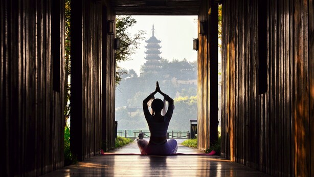 Una persona en posición meditativa en un corredor con arquitectura tradicional y una pagoda al fondo.