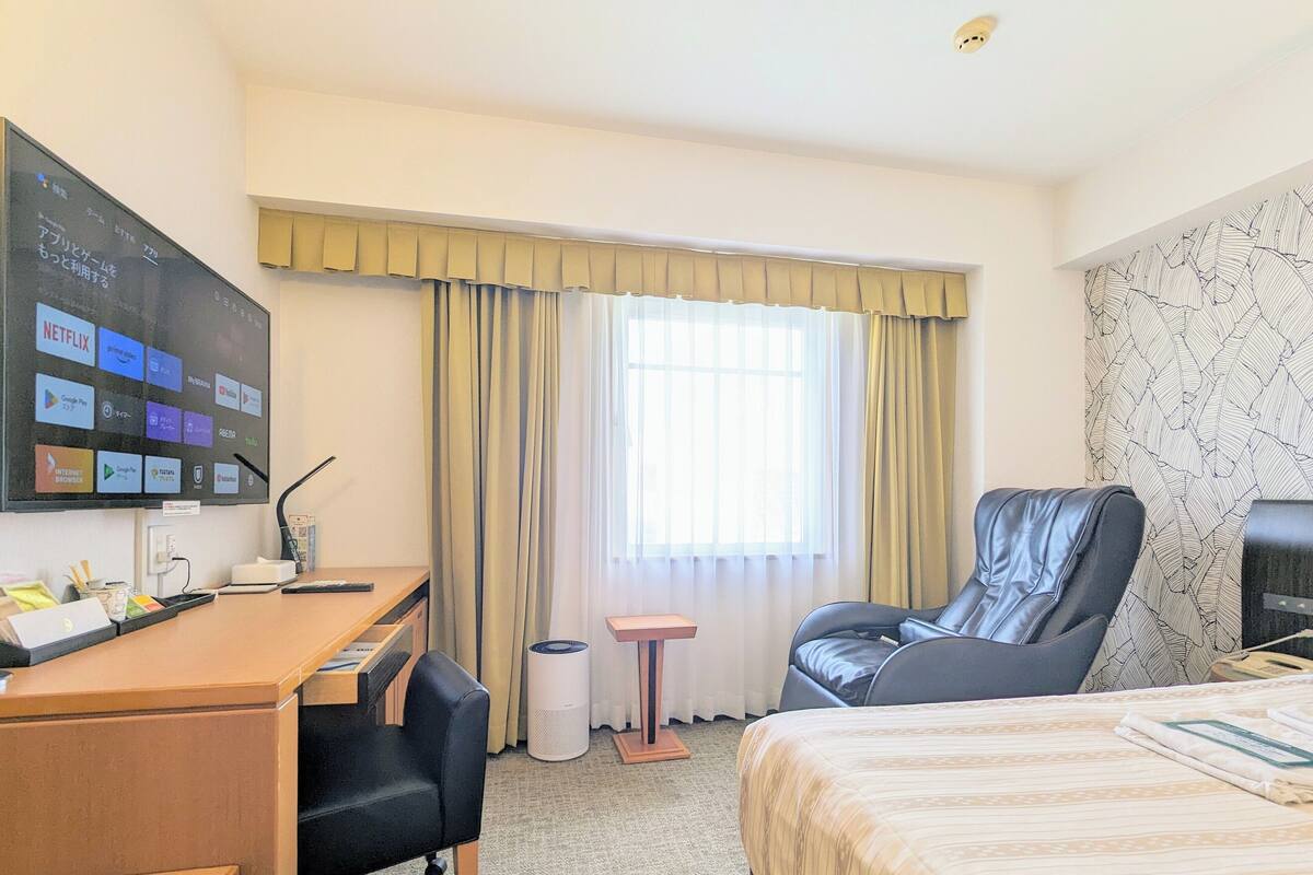Une chambre d’hôtel équipée d’un bureau, d’une chaise, d’un lit, d’une télévision et d’une fenêtre avec des rideaux.