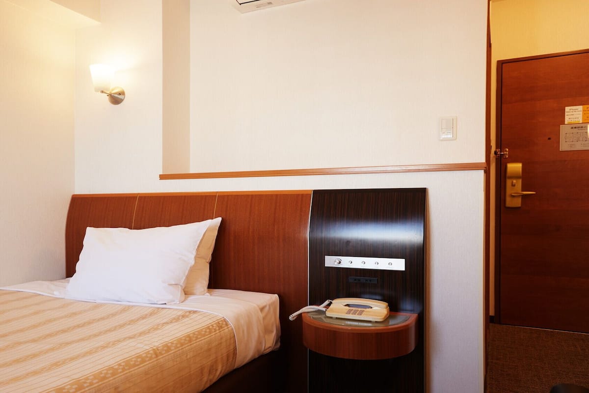 A hotel room with a bed, a bedside telephone, and a wall-mounted light fixture.