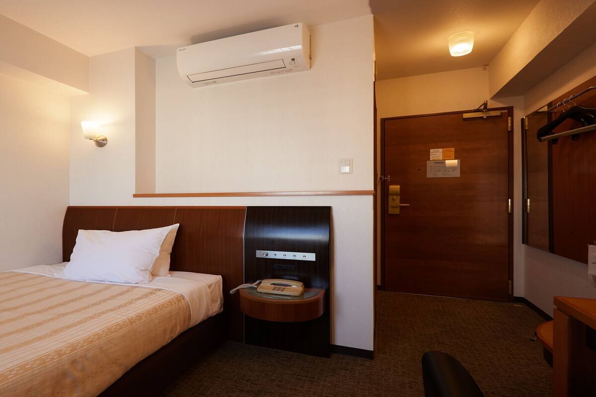 A hotel room with a bed, a bedside telephone, an air conditioning unit, and a wooden door.