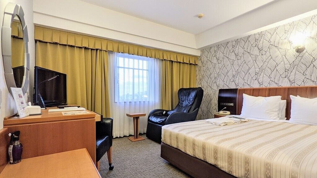 A hotel room with a bed, a desk with a television, a chair, a bedside table, and a window with curtains.