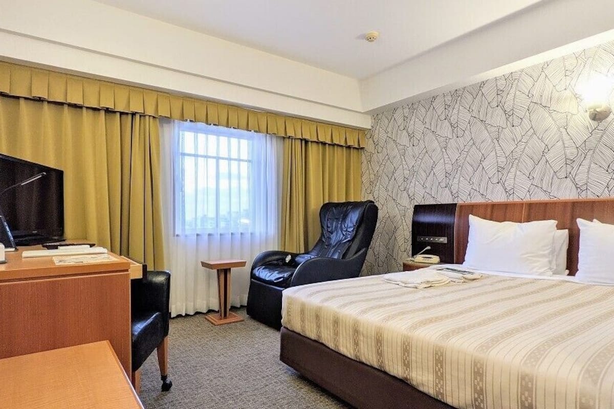 A hotel room with a bed, a desk with a television, a chair, a bedside table, and a window with curtains.
