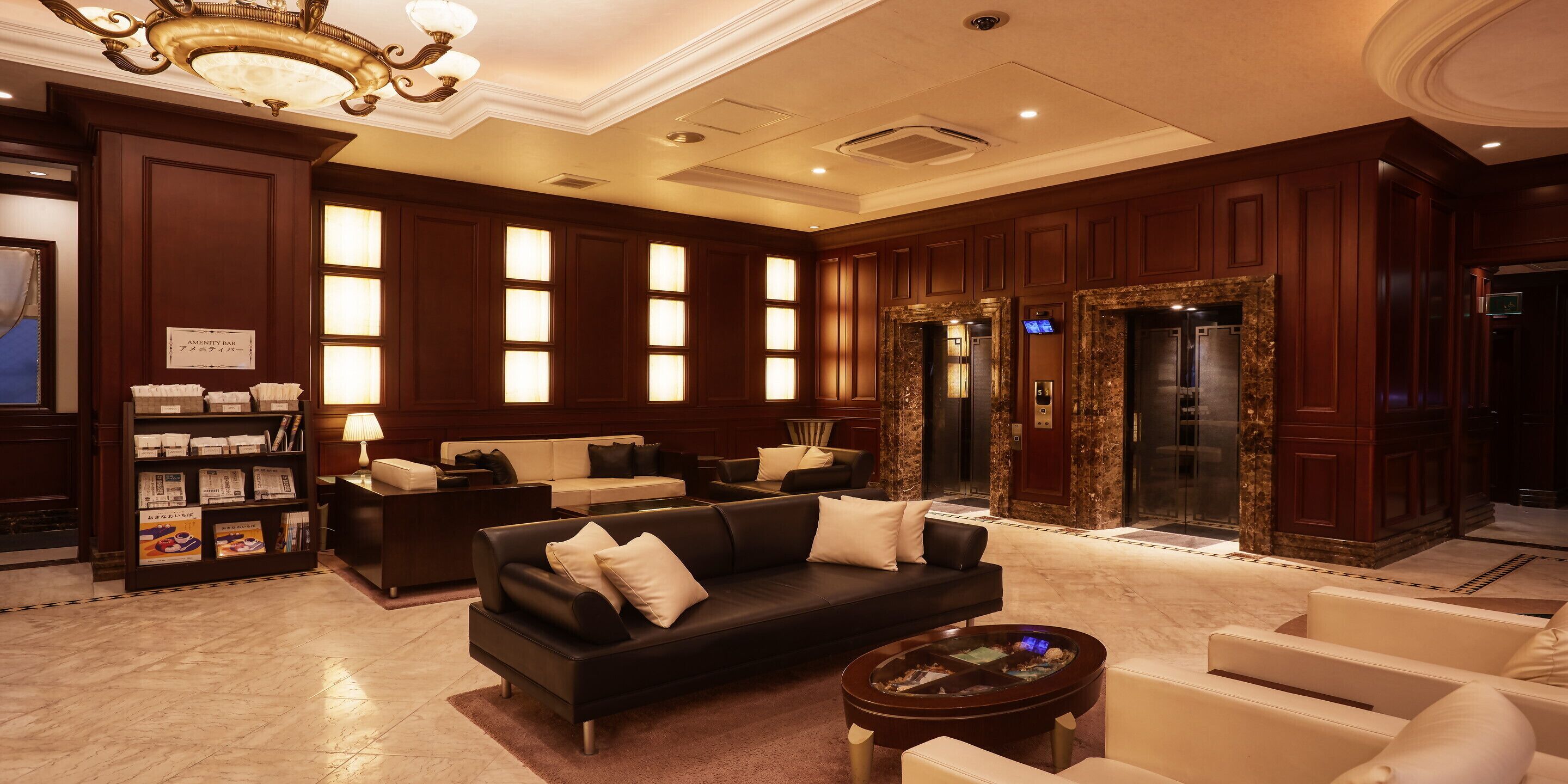 A spacious lobby with dark wood paneling, a black leather sofa, white armchairs, a marble floor, and a reception desk with a sign reading "AMENITY BAR".