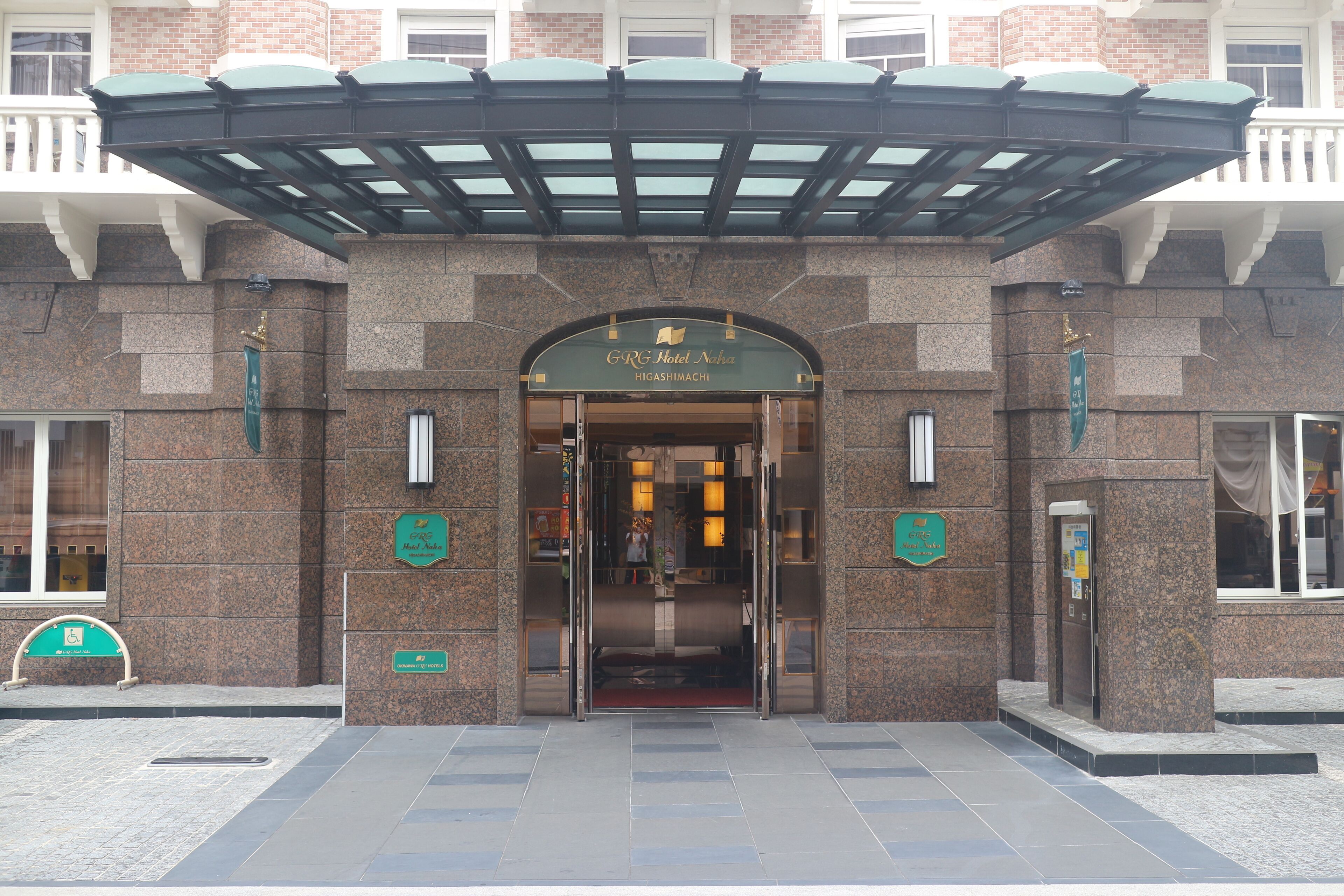 La entrada del Hotel G.R.G Naka, con un vestíbulo cubierto, fachada de piedra y un cartel que indica accesibilidad para personas con discapacidad.