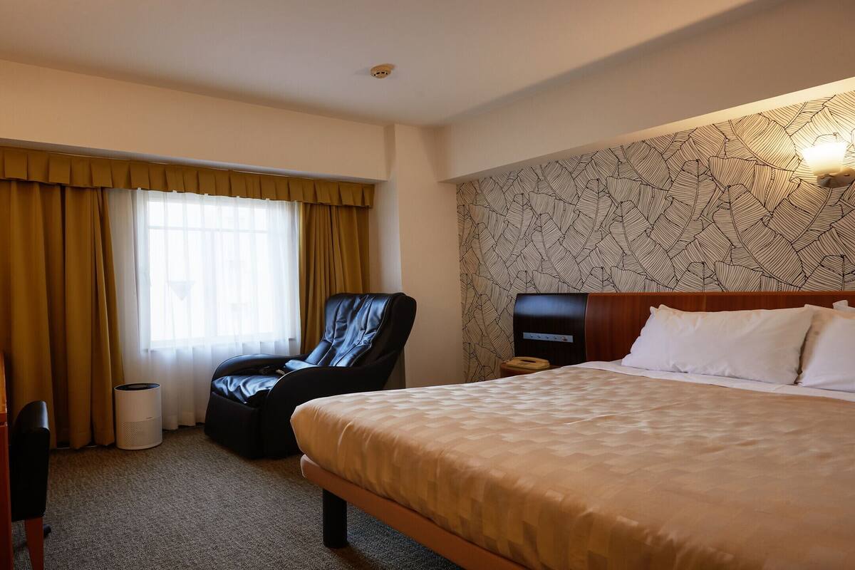 A hotel room with a bed, a desk with a computer, a chair, and a large window with curtains.