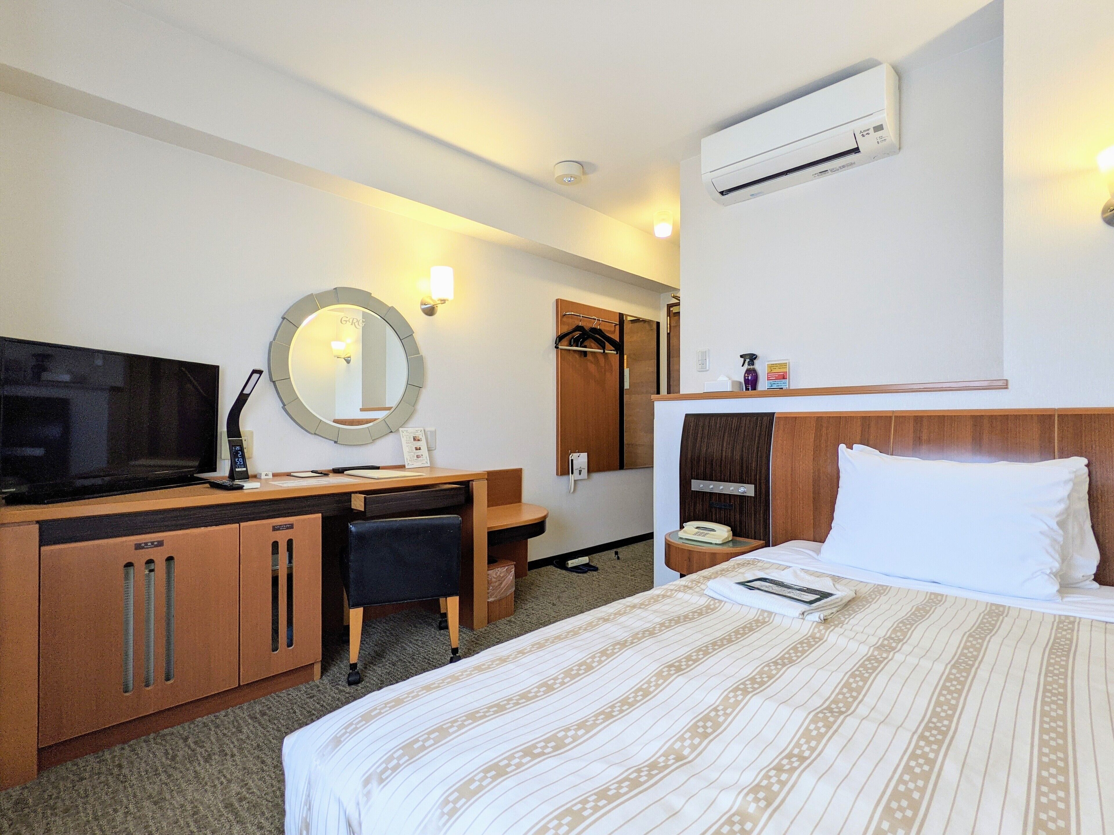 A hotel room with a bed, a desk with a TV, a mirror, an air conditioner, and a bedside table.