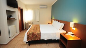 Une chambre d’hôtel comprenant un lit, une table de chevet avec une lampe, un téléviseur fixé au mur, un petit réfrigérateur et une fenêtre avec des rideaux.