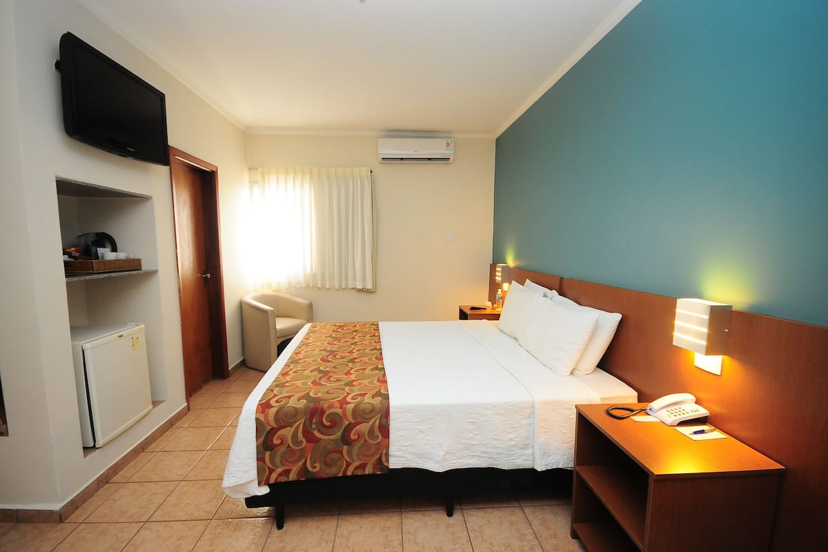 A hotel room with a bed, bedside table with a lamp, a TV mounted on the wall, a small refrigerator, and a window with curtains.