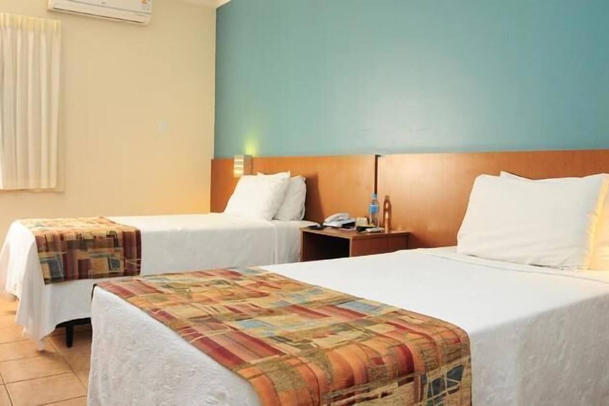 A hotel room with two beds, a wooden headboard, a nightstand with a phone and a bottle, and a window with curtains.