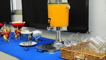 A juice dispenser with a blue tablecloth, a basket of clear glasses, and a bowl of fruit skewers.
