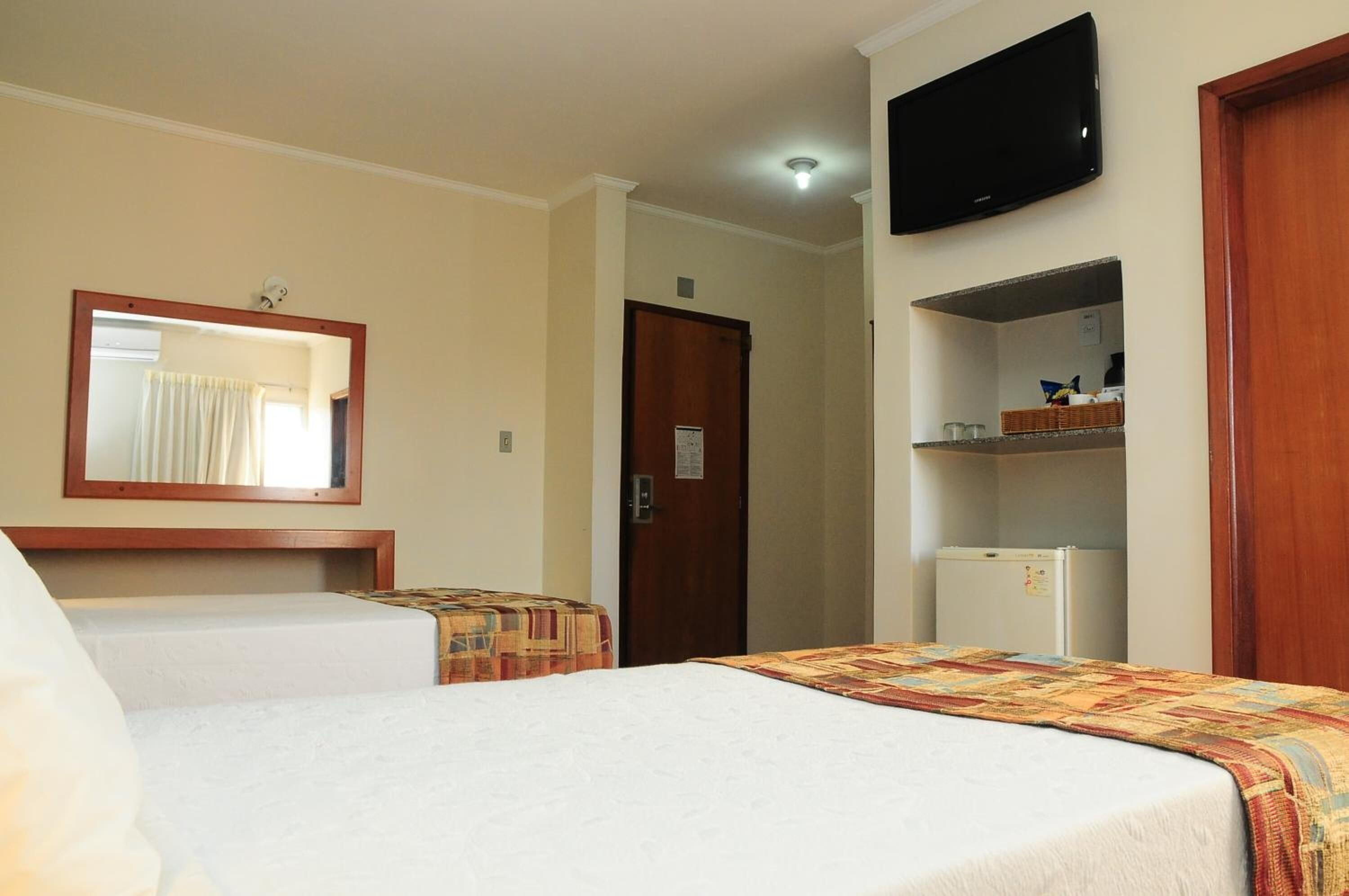 A hotel room with two beds, a television, a mirror, a door, and a small refrigerator.